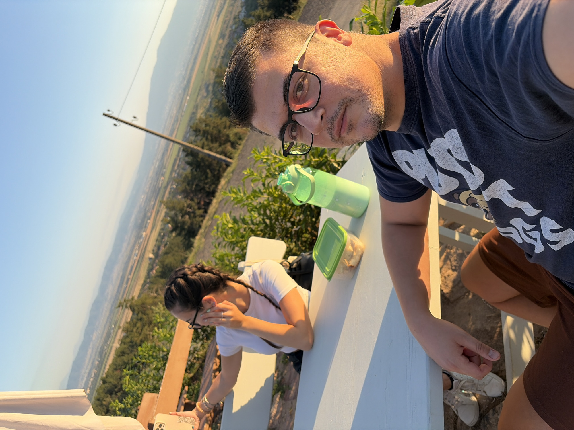 In Sofia, Bulgaria, a young man and woman are seated at a white outdoor table, likely on a hill overlooking the city. The man, in the foreground, is taking a selfie, his face turned towards the camera. He wears glasses and a dark t-shirt. The woman, in the background, is also looking at her phone, which she holds in her left hand. She has braided hair, wears glasses and a white t-shirt, and rests her right hand on her face. The table holds a green water bottle and a plastic container with food. The background shows a scenic view of a valley with agricultural fields and distant mountains. The lighting suggests it is late afternoon or early evening, with the sun casting a warm glow on the scene.