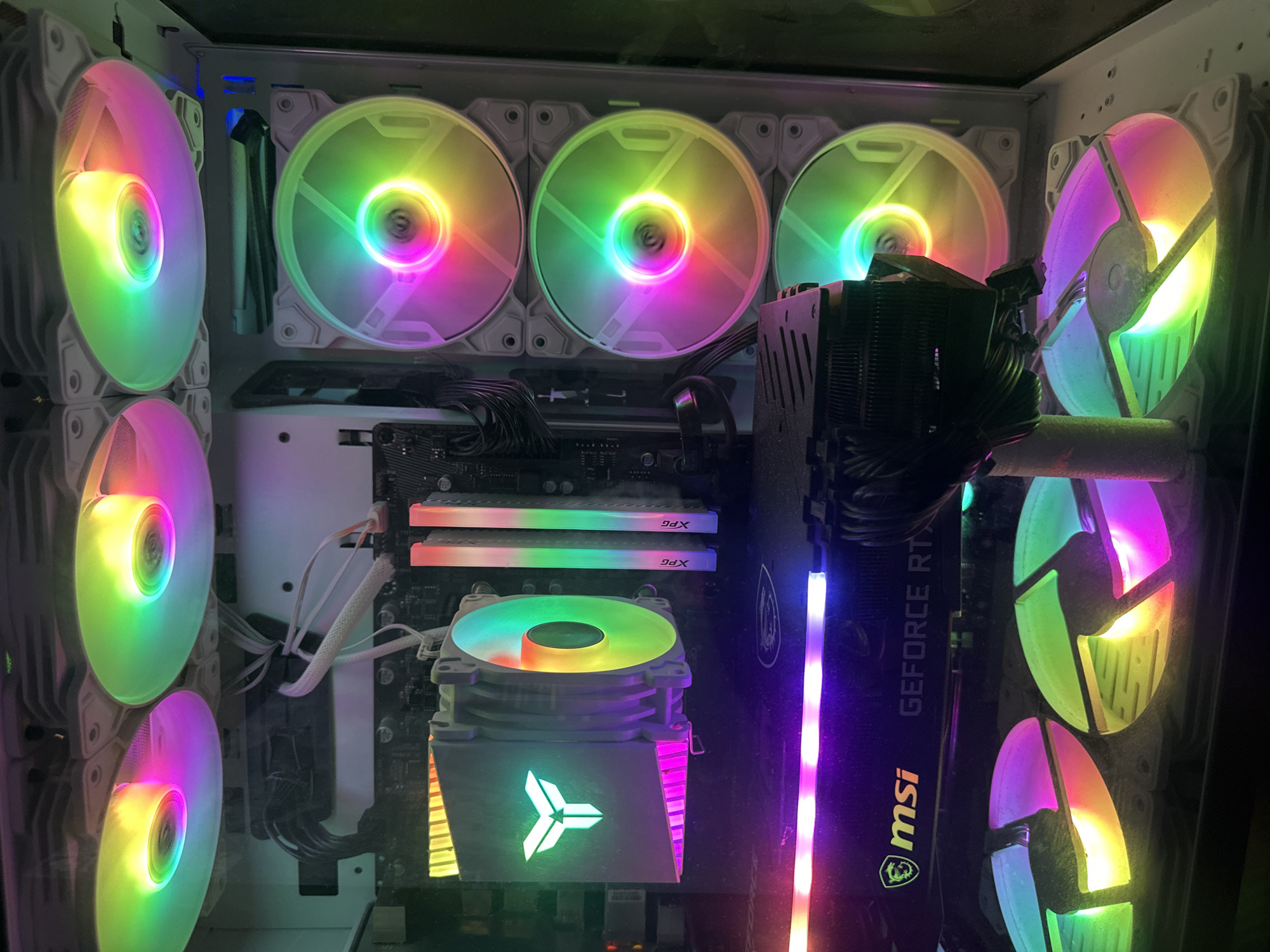 This media file showcases the interior of a computer, highlighting its components and RGB lighting. The primary subjects are the various computer parts, including several fans, RAM sticks, a CPU cooler, and a graphics card. The scene is set within a computer case, which appears to be a white ATX tower.
The computer is powered on, as evidenced by the illuminated RGB lighting on the fans and other components. The fans, which are arranged on the side and front of the case, are displaying a vibrant rainbow color spectrum. The RAM sticks are also lit up with a similar colorful glow, as is the CPU cooler, which features a prominent logo illuminated with green and pink light. The graphics card, identified by the text "GEFORCE RT" and the MSI logo, also has some subtle lighting along its edge.
Notable details include the dust accumulation on some of the components and fans, suggesting the computer has been in use for some time. The wires within the case are neatly managed, contributing to a clean internal aesthetic despite the dust. The lighting creates a visually striking, high-tech appearance. There is no discernible time of day or weather context. The overall emotion evoked is one of advanced technology and customization.
Visible text includes "GEFORCE RT" and the "msi" logo on the graphics card, as well as "XPG" on the RAM sticks. The CPU cooler also displays a geometric logo.