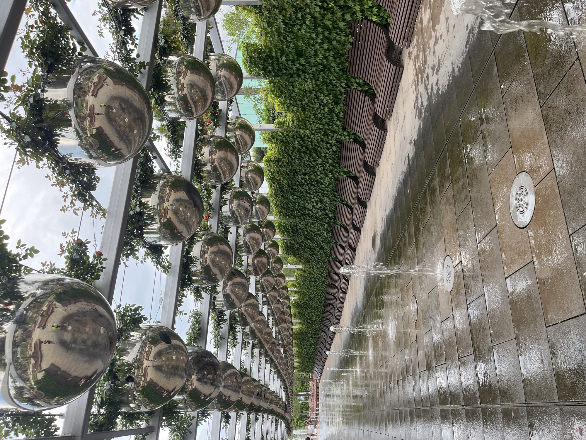 The image captures a park scene in Krasnodar, Russia. Above, a canopy structure supports numerous reflective spheres interspersed with vegetation, possibly roses. Adjacent to this structure runs a vertical wall of green plants. The ground is paved with large tiles, and a linear water feature consists of several small geyser-like fountains that spray water onto the tiles. The pavement appears wet. At the far end of the park, blurred human figures are visible. The image is taken from an angle that emphasizes the length of the park.