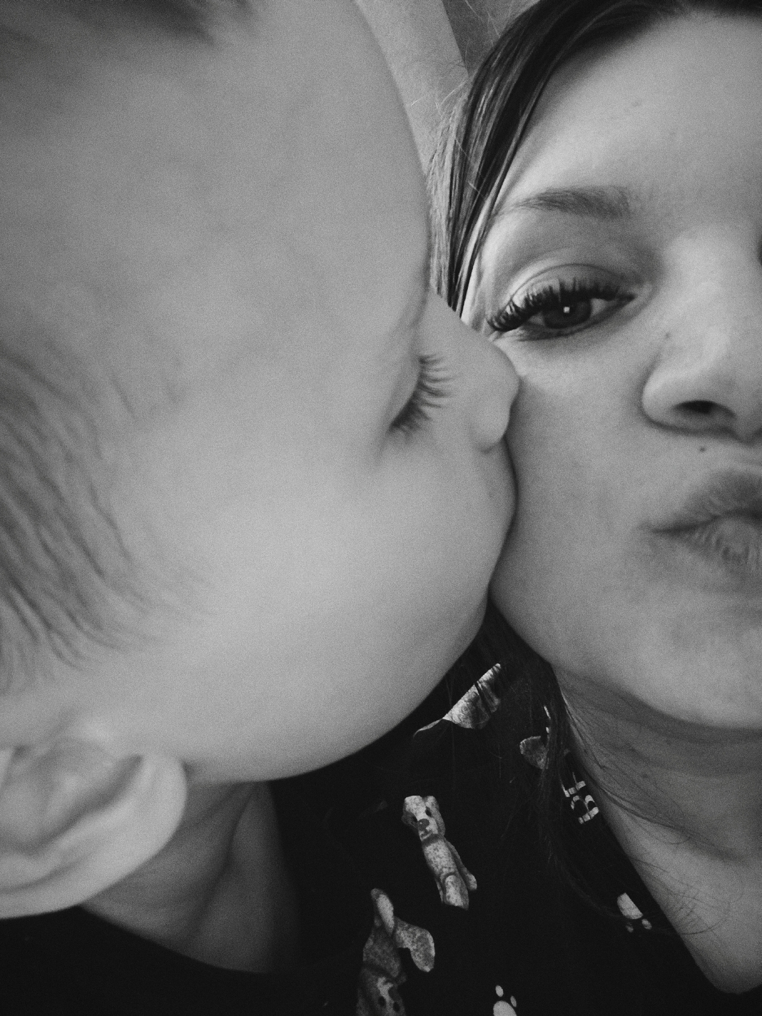 A black and white, medium close-up captures a mother and child interacting. The child, positioned on the left, is kissing the mother's cheek. The child has fine hair, visible eyelashes, and a soft cheek pressed against the mother's face. The mother is positioned on the right, with her face partially visible. Her eye is clearly defined, and she has long eyelashes. Her lips are pursed as if returning the kiss. The mother is wearing a dark shirt with a pattern of small figures on it. The background is indistinct. The location is not discernible from the image.