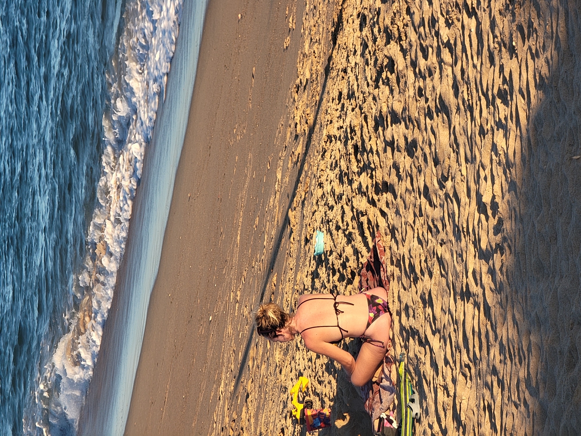 In Quarteira, Portugal, a woman sits on a towel on a sandy beach, facing the ocean. She is wearing a dark bikini with a colorful floral pattern. Her hair is pulled up in a bun, and sunglasses are resting on her head. In front of her, a few beach toys, including a yellow shovel and a bucket, are visible. The ocean water is blue with white waves gently rolling onto the shore. The sand is textured with rippled patterns from the wind and footprints, and long shadows are cast across it from an unseen light source, suggesting it is either early morning or late afternoon.
