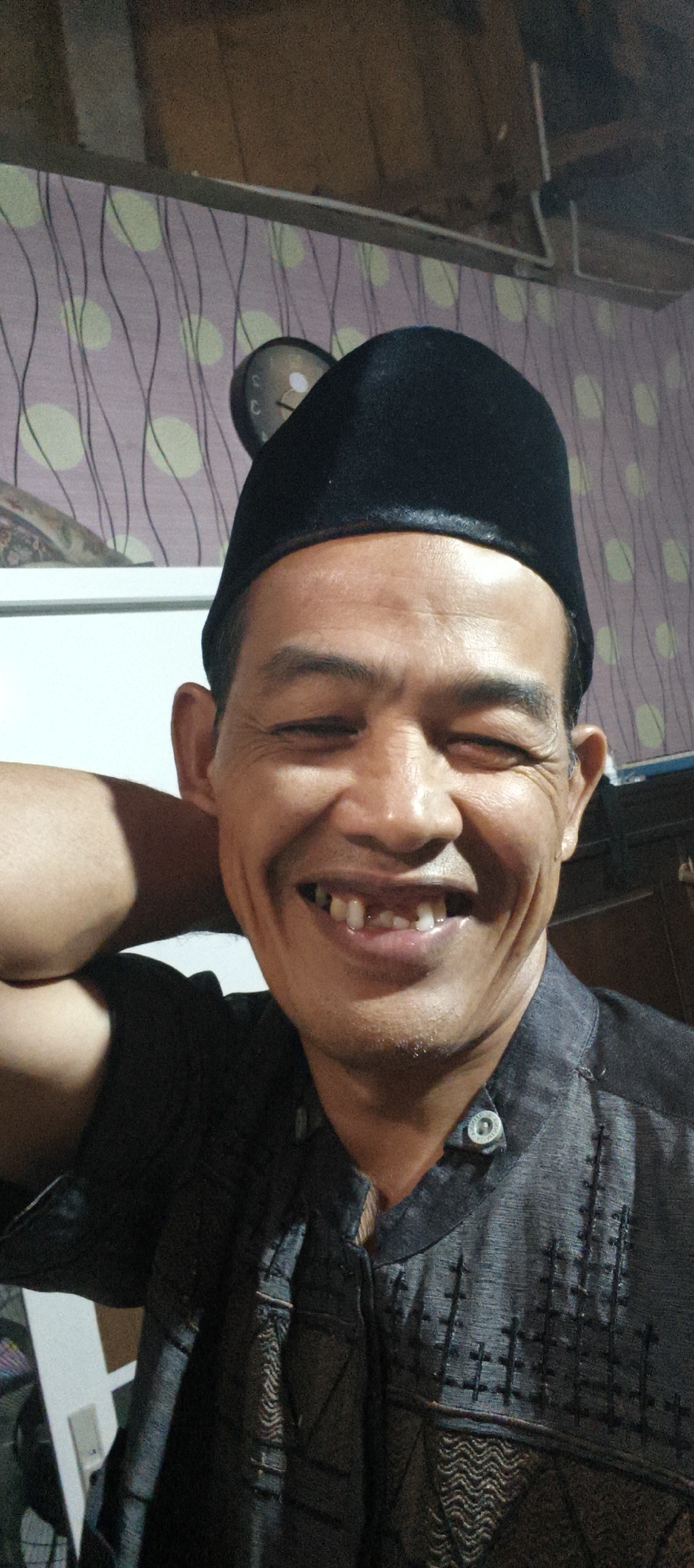 A medium shot shows a man with his left arm behind his head, smiling at the camera. He is wearing a black peci hat and a dark grey shirt with a patterned design and a button at the collar. The man's teeth are visible in his smile, with some missing. The background is a wall with a light purple wallpaper featuring a pattern of wavy dark lines and light green circles. A clock with a black face and white hands is visible on the wall behind the man's head. The setting appears to be indoors, likely a residential room in Muaro Jambi, Indonesia.