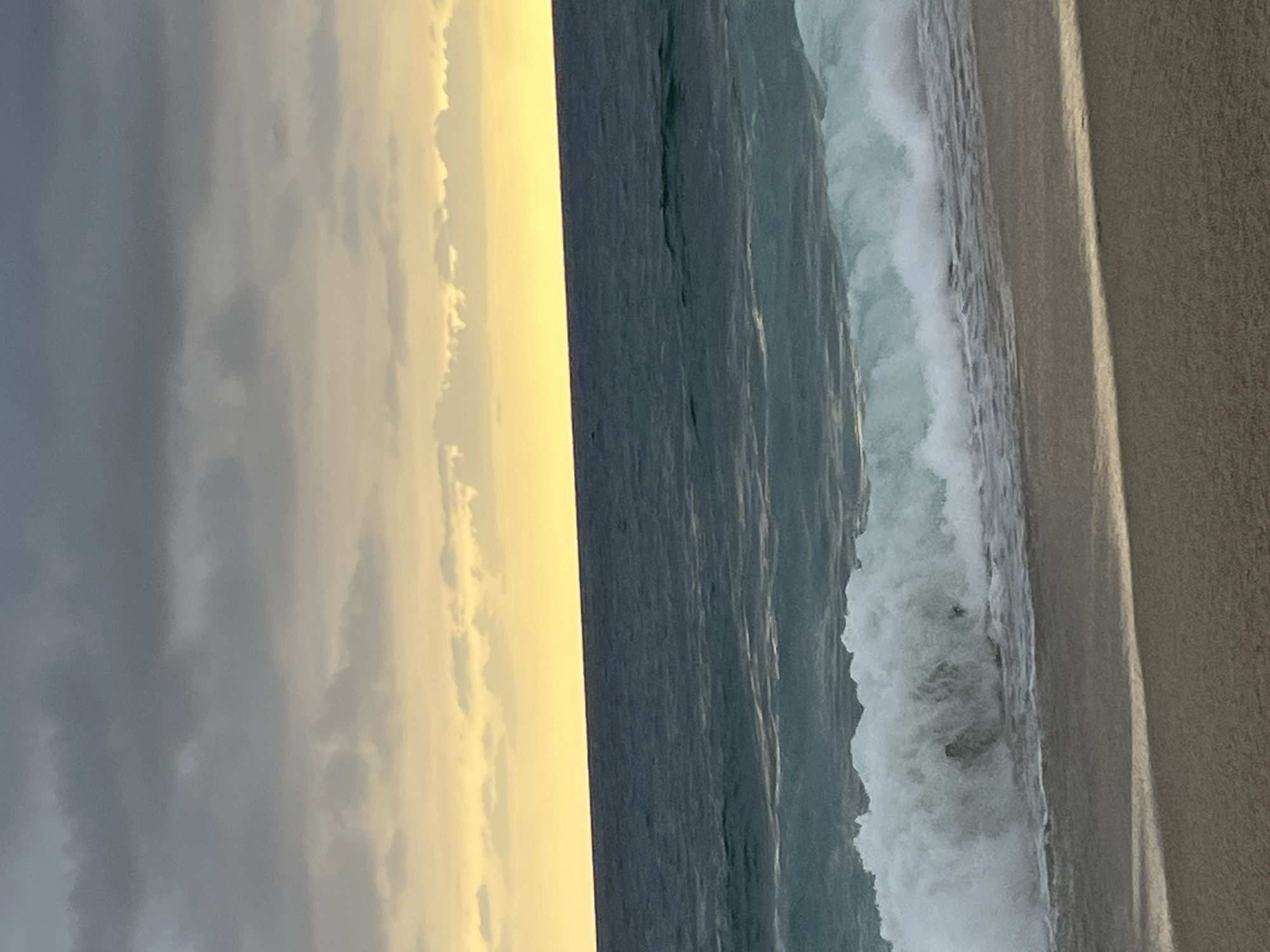 The image shows a beach scene, possibly near Alanya, Turkey, featuring a sandy beach on the right, with foamy waves crashing onto it. The water transitions from a dark gray-blue in the center to white foam near the shore. The sky on the left side of the image is a mix of gray and yellow hues, likely indicating either a cloudy or possibly a sunrise/sunset scene. The location is inferred due to the beach setting in Alanya, Turkey.