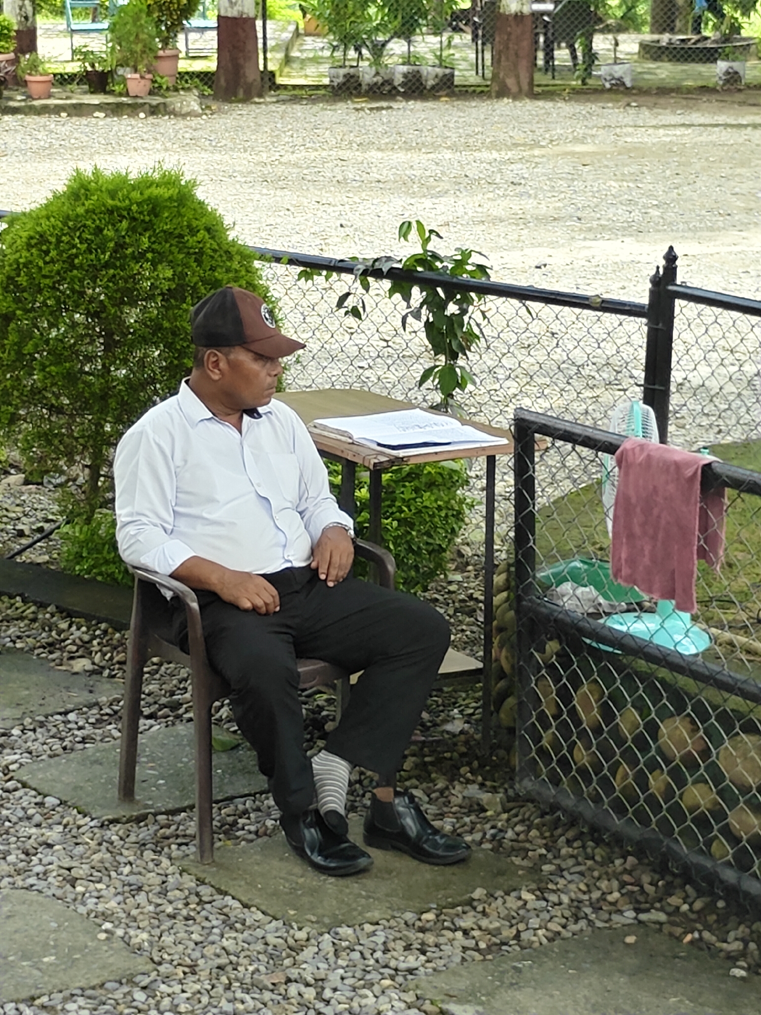 A man wearing a white long-sleeved shirt, black pants, a brown and black baseball cap, and black shoes with striped socks is sitting in a brown plastic chair. He is looking to his right. To his left is a small green bush. Behind him is a chain-link fence with a maroon towel draped over it and a small, blue electric fan. A table with papers is situated to his right, and beyond that is a gravel-covered ground with some plants and trees in the background. The scene appears to be outdoors.