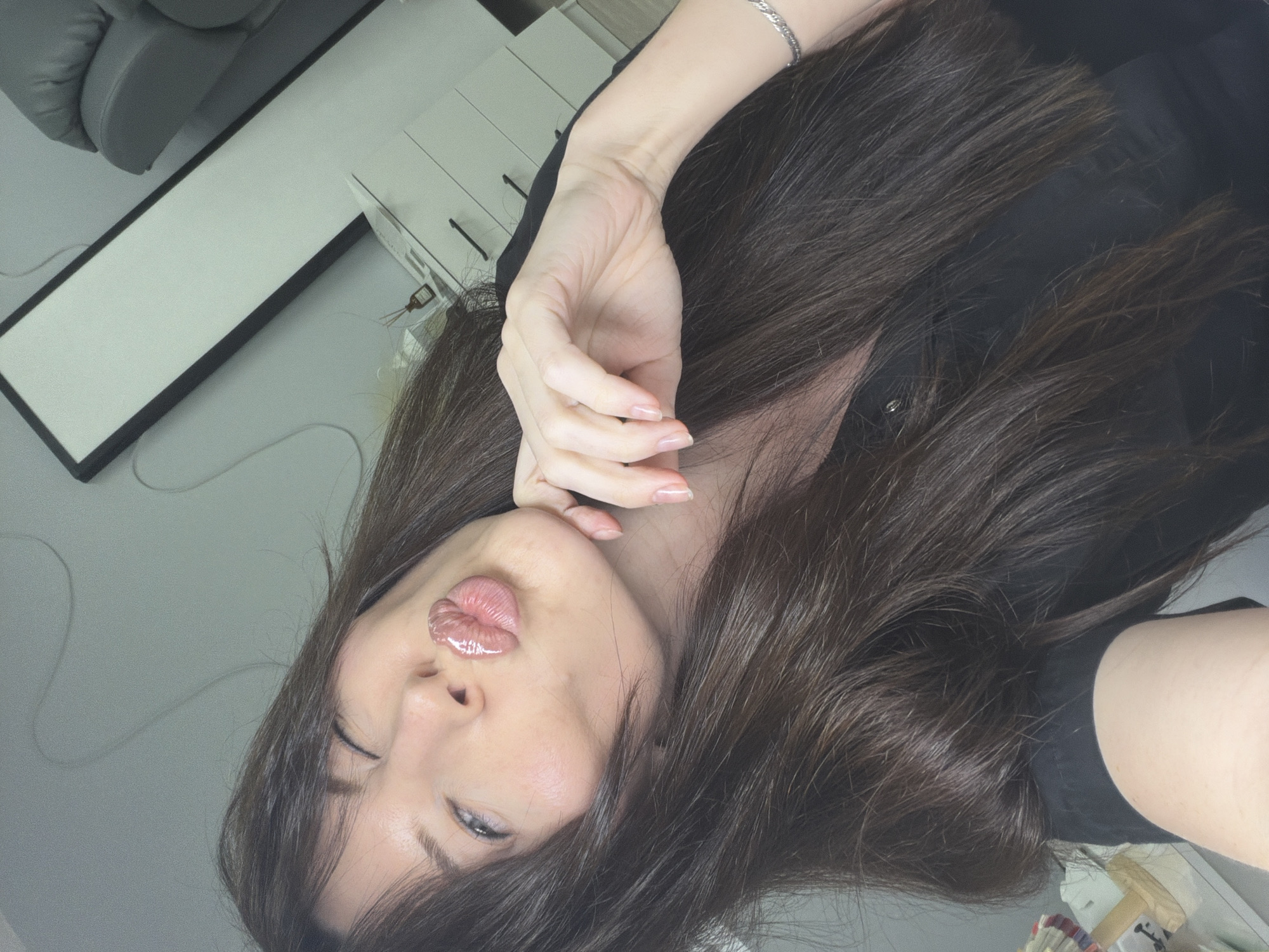 A woman with long, dark brown hair, partially covering her left eye, is posing indoors. She has her right hand raised near her cheek and glossy lips. She is wearing a dark-colored shirt. A grey chair and a white table or cabinet are visible in the background. There is no clear indication of the location being Perm, Russia.