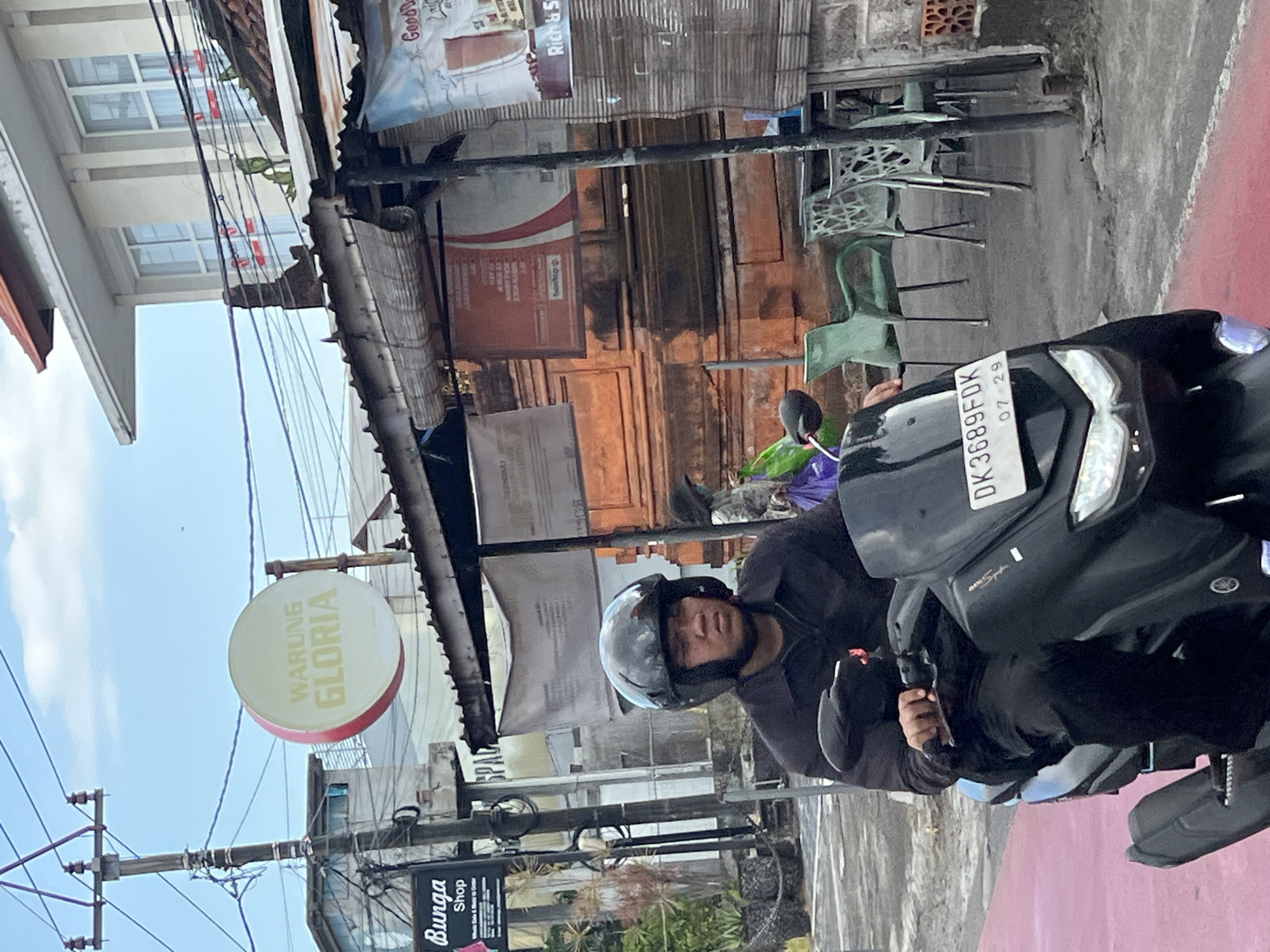 The image depicts a man on a motorcycle in North Kuta, Indonesia. The motorcycle has a license plate reading "DK 3689 FDK" with an expiration date of "07-29." The man is wearing a dark-colored jacket and a black helmet. Behind him, there is a sign for "Warung Gloria," and a sign for "Bunga Shop." The streetscape includes power lines, buildings with weathered facades, and outdoor seating. The environment appears to be a commercial or residential area with a mix of shops and eateries.