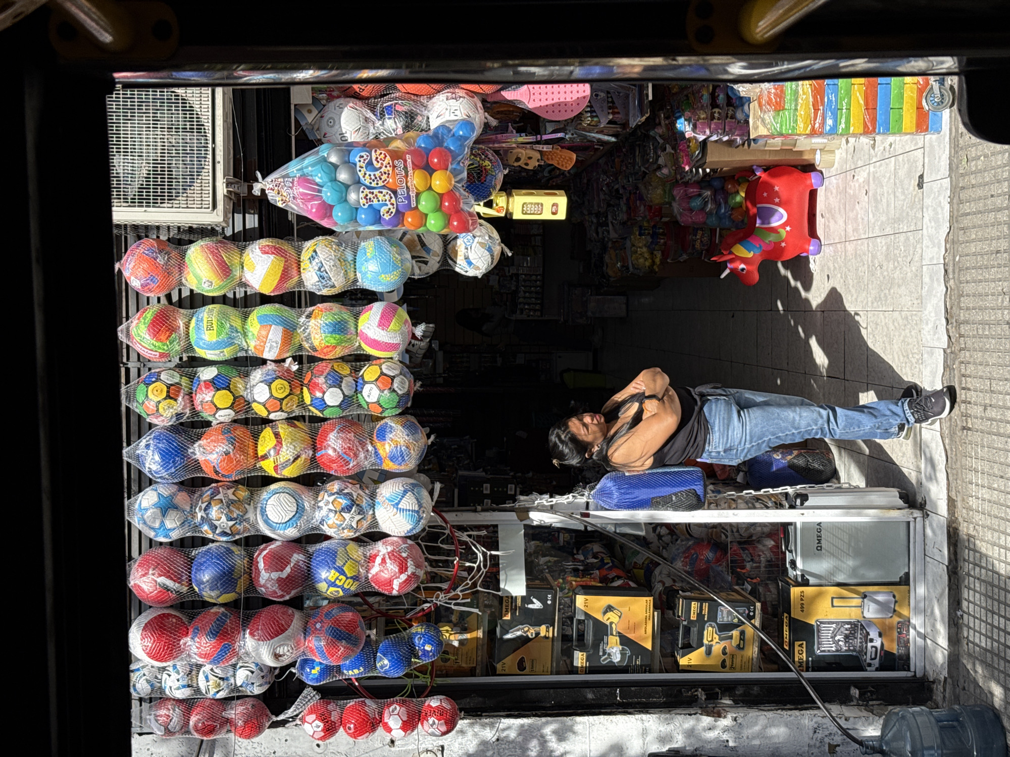 A street-level view shows a small shop in Buenos Aires, Argentina, as seen from inside a passing vehicle. The shop’s exterior is cluttered with goods: rows of colorful soccer balls and other toys hang near the top of the storefront. Various toolboxes, cases, and equipment are displayed on shelves at the bottom. A person is lying on a makeshift bed on top of the shelves, using a blue bag as a pillow. The scene is lit by sunlight, casting shadows on the sidewalk in front of the shop. The framing of the shot, partially blocked by the vehicle's interior, suggests a snapshot taken quickly while passing by.