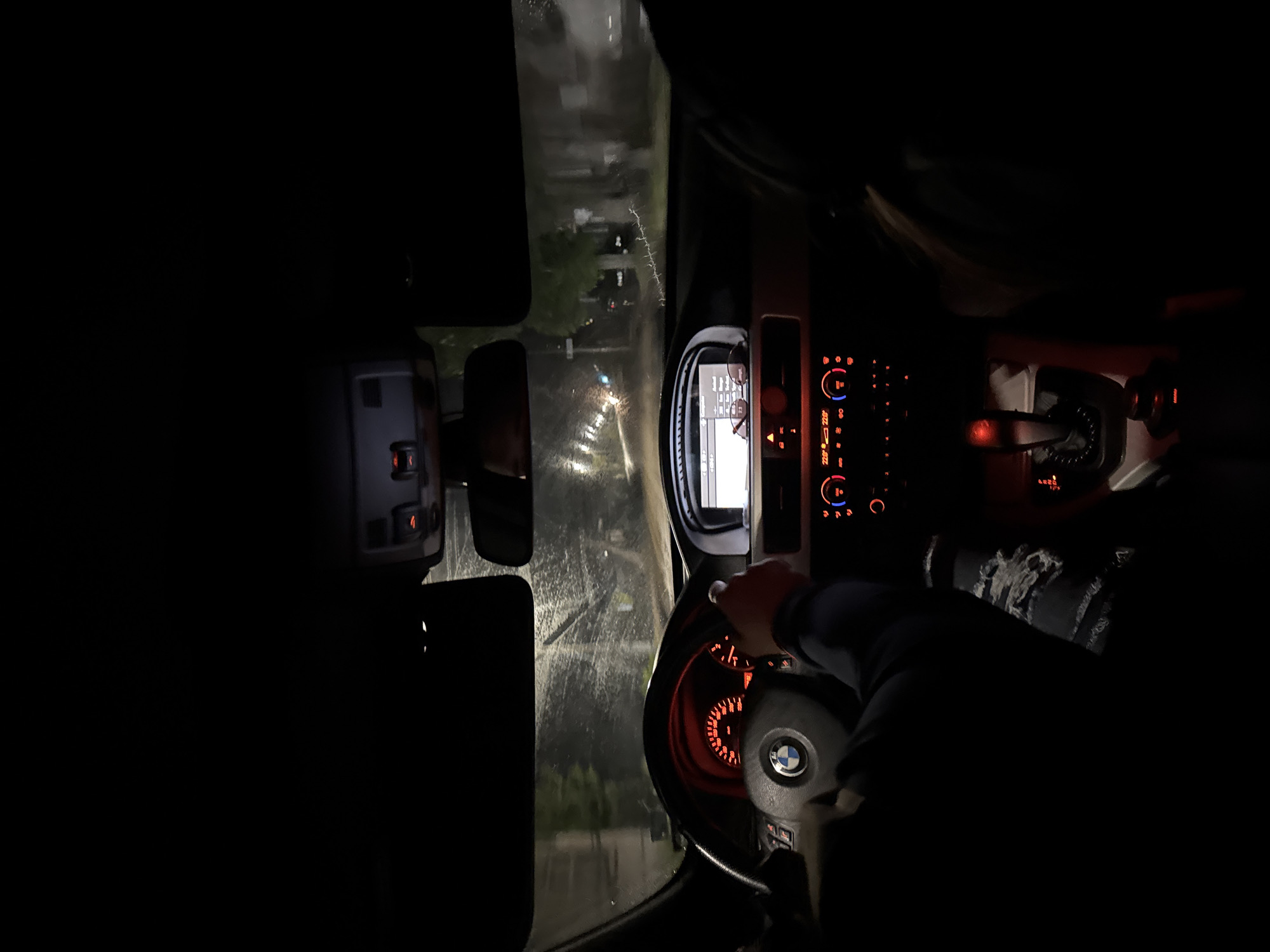 A nighttime view from inside a vehicle, likely a BMW based on the steering wheel emblem. The dashboard is illuminated with orange and red lights. A person's hand is visible on the steering wheel. Rain streaks are visible on the windshield, and streetlights and houses are visible in the background. The location is unknown and cannot be determined from the image, therefore the specific city and country cannot be identified.