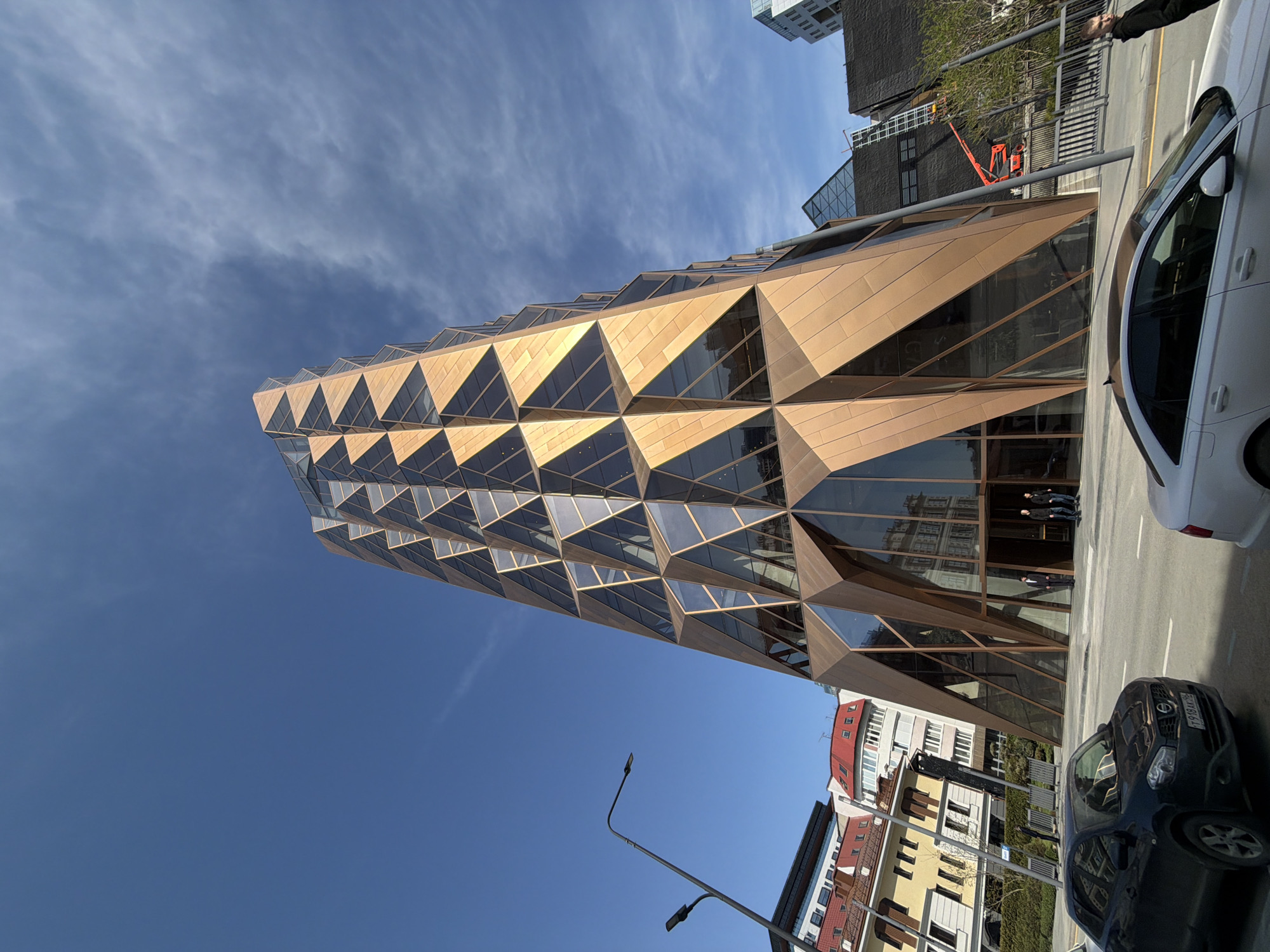 The image captures a modern architectural structure in Yekaterinburg, Russia, known as the "Anglian Building." The building has a distinctive design, featuring a facade composed of numerous triangular panels, some of which are glass, reflecting the surroundings. The panels are predominantly golden in color.
The angle of the shot emphasizes the building's height and unusual shape. It is situated among other buildings of various styles, including older, traditionally styled buildings with red roofs. The sky is visible in the background, partly cloudy with a blue hue.
A street scene is captured at the base of the building, where cars are parked, including a white sedan and a dark-colored SUV. A street lamp post is also present.