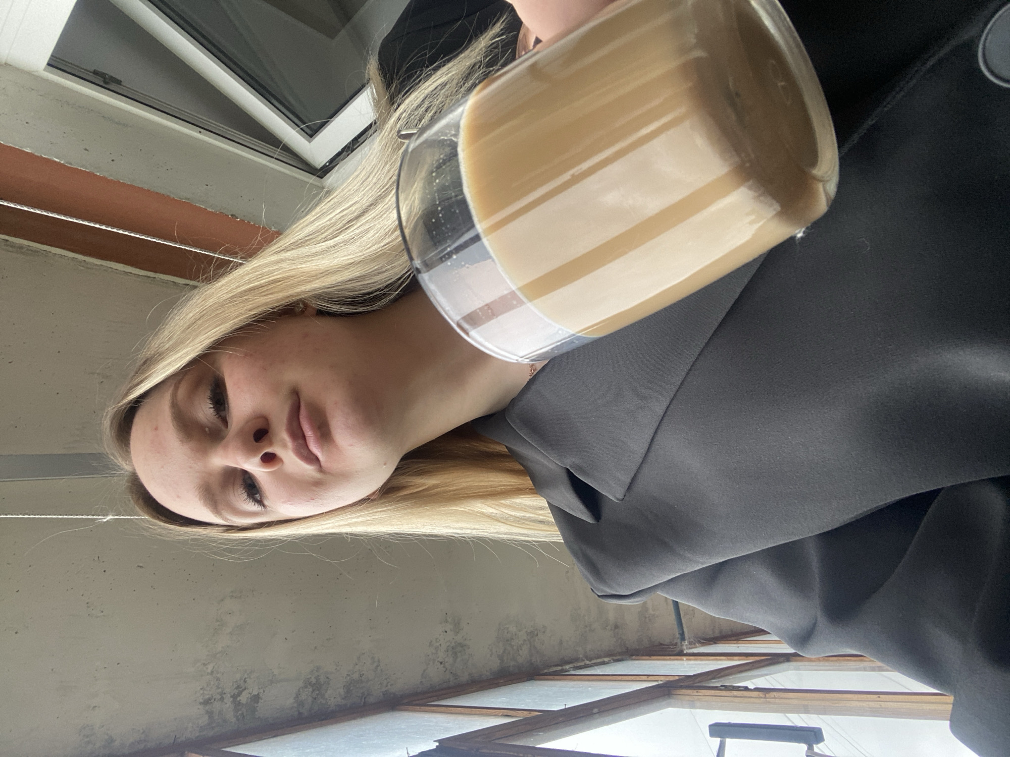 A young woman with long, straight blonde hair and fair complexion is depicted in a low-angle, eye-level shot, looking directly at the camera. She has visible minor blemishes on her forehead and chin. She wears a dark charcoal grey blazer with a structured lapel. A subtle rose gold-toned necklace is visible around her neck. Her right hand, partially visible, holds a clear glass filled with a milky, light brown beverage, consistent with coffee or tea with milk, nearly to the brim.
The background features a textured, light grey concrete ceiling with faint discoloration and dust. To the left, a vertical window or glass panel with a dark frame is visible. To the right, a white-framed window opens to an indistinct, light-toned exterior. An orange-brown horizontal beam or pipe runs above the right window, against a lighter background. The scene appears to be an indoor or sheltered outdoor space within a building in Unknown, Unknown.