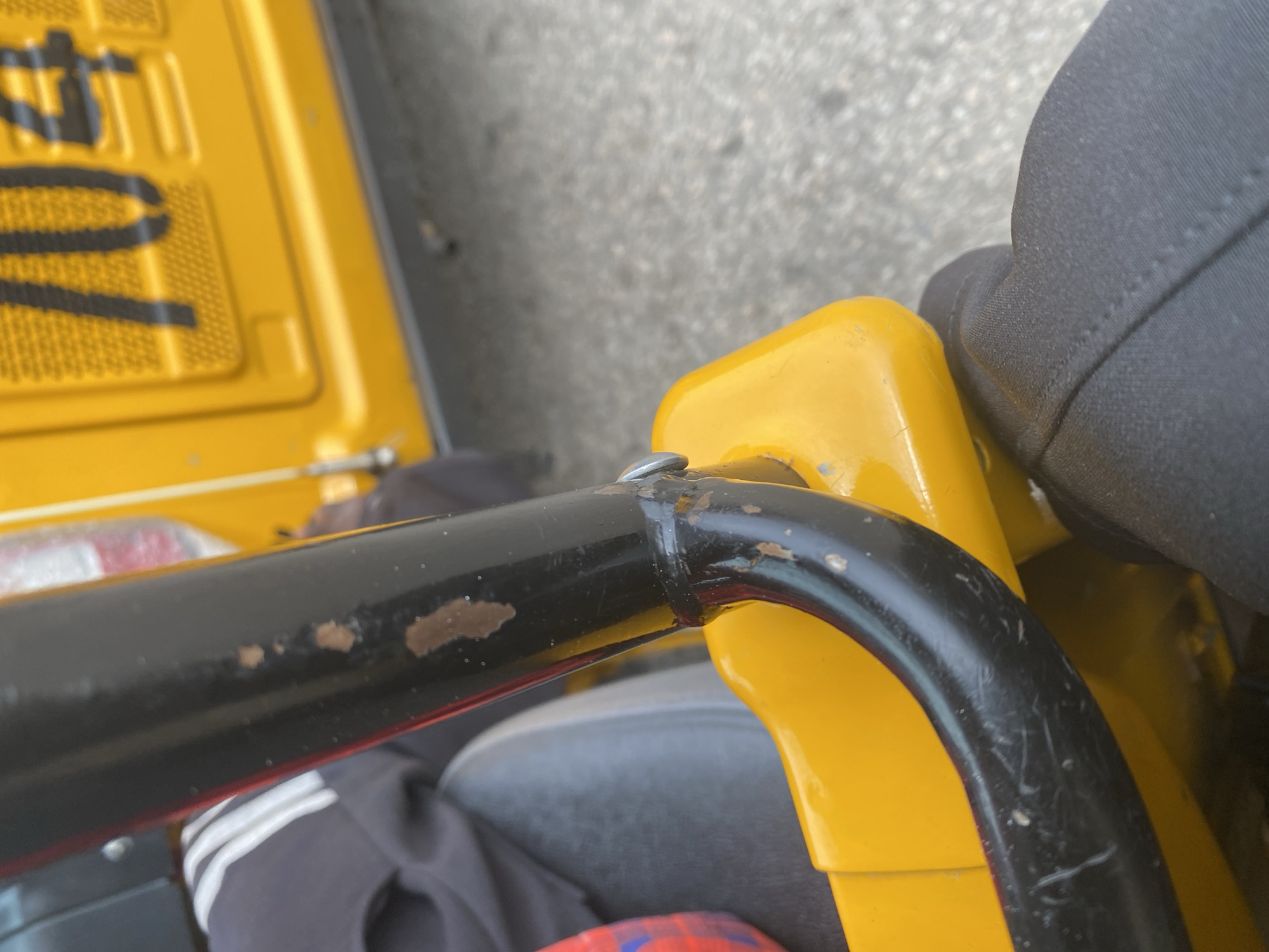 The image shows a close-up view of a yellow object, possibly a vehicle or equipment, with a black metal bar attached to it. The metal bar has visible signs of rust and scratches. A person's dark clothing, likely pants, is visible on the right side of the frame, suggesting they are sitting on or near the yellow object. In the background, a grey, paved surface can be seen, indicating an outdoor setting. The lighting suggests it is daytime. There is no discernible text in the image. The scene appears to be in Jalingo, Nigeria, based on the provided location context. The overall impression is one of everyday use, with the wear and tear on the metal bar indicating the object is functional but not new.