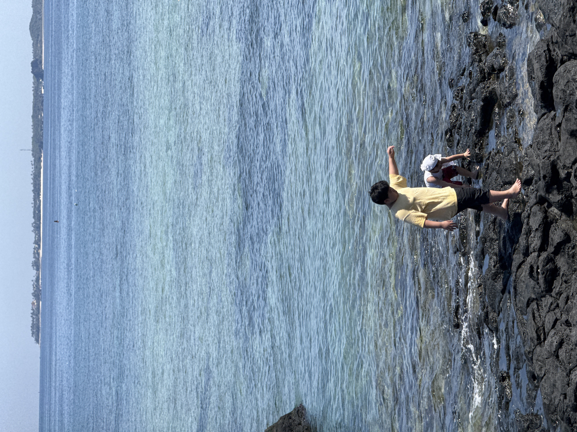 Two children are standing on a rocky shoreline with the ocean in the background. The child in the foreground, on the left, is wearing a yellow t-shirt and black shorts. They are barefoot and appear to be looking down towards the water. The second child, in the middle ground on the right, is wearing a white hat and red shorts. They are crouched down and also appear to be looking at something in the water or on the rocks. The water is a clear blue and the shore is made up of dark, wet rocks. In the distance, a line of trees and buildings is visible on a peninsula, suggesting a coastal town or island. The sky is a pale blue and appears to be clear. The scene captures a moment of exploration and play by the sea.
