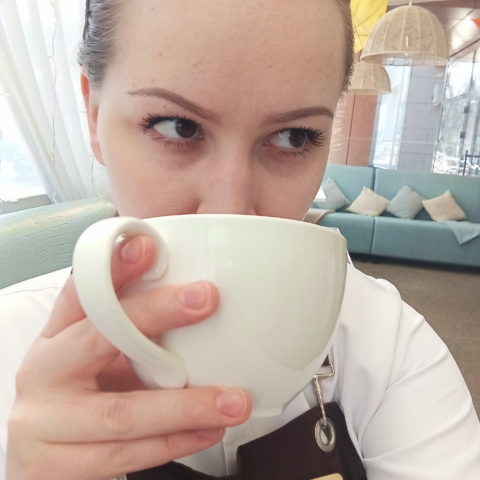A young woman is holding a large white mug in front of her face, partially obscuring her mouth and nose. She is looking to her right. Her brown eyes are visible above the mug's rim. She has neatly groomed eyebrows and noticeable mascara. Her hair is pulled back and away from her face. She is wearing a white collared shirt and a dark brown apron. The metal clip and grommet of the apron are visible near the mug. Behind her, there is a light blue upholstered sofa with matching cushions. In the upper background, there are woven light fixtures, and beyond the windows, there are hints of an outdoor scene, possibly with snow or light-colored buildings. The location is possibly in a café or restaurant in the city of Chaikovsky, Russia.