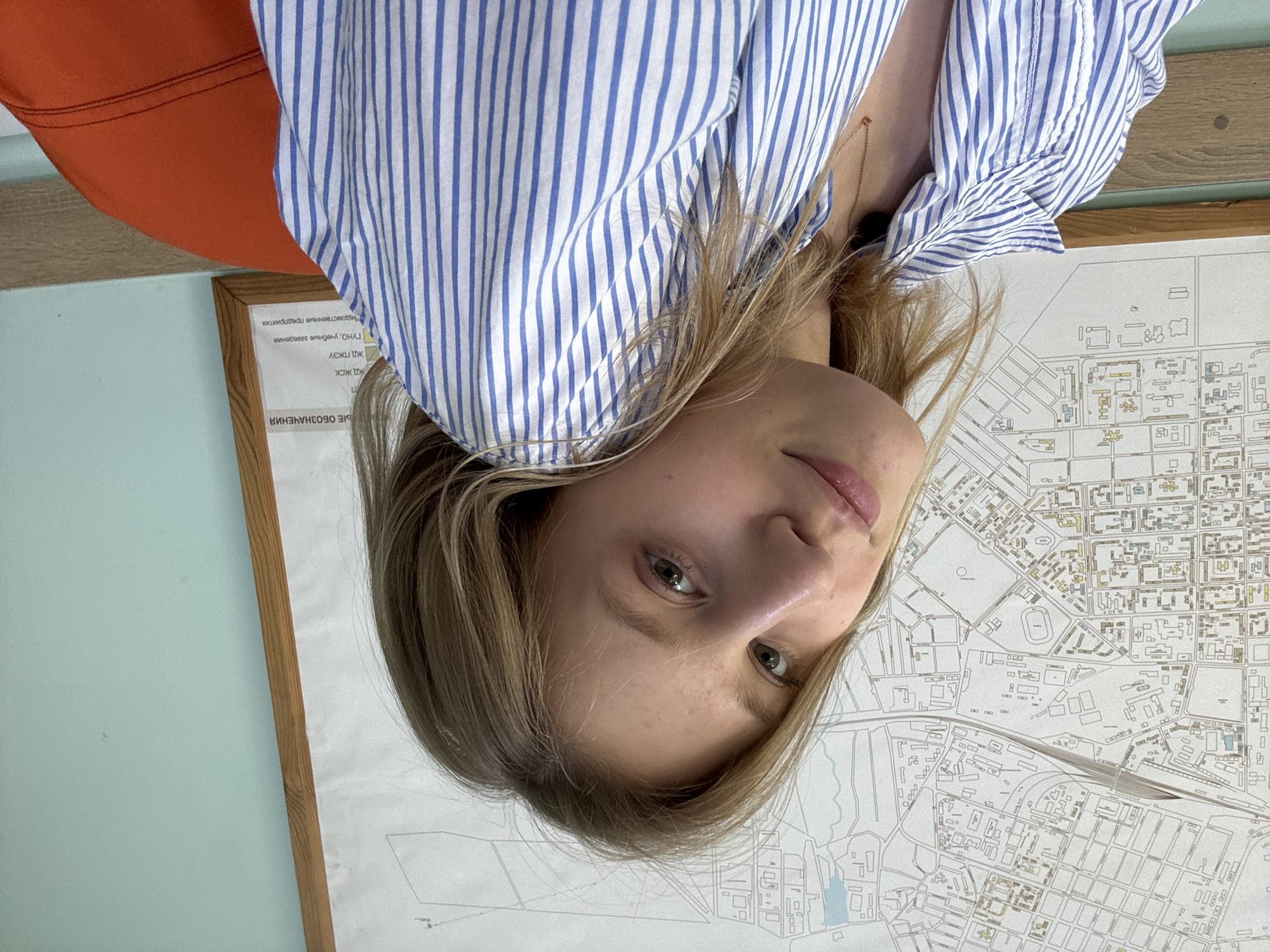 In this close-up, indoor shot, a young woman with blonde hair, wearing a blue and white striped collared shirt and a delicate gold necklace, is positioned at a slight angle, looking towards the camera. Her head is tilted, with her hair cascading over her shoulder. Behind her, a large, detailed map hangs on a light blue wall, framed by a wooden border. The map appears to be a city plan, featuring intricate lines representing streets, buildings, and other urban features. Some text in Cyrillic script is visible on a section of the map that is slightly out of focus. A portion of an orange chair or cushion is visible in the lower right corner of the frame. The lighting is soft, suggesting an indoor environment, possibly an office or classroom, given the presence of the map. The Tiraspol Fortress, a prominent landmark in Tiraspol, Moldova, might be depicted on the map, but it is not explicitly identifiable from this perspective.