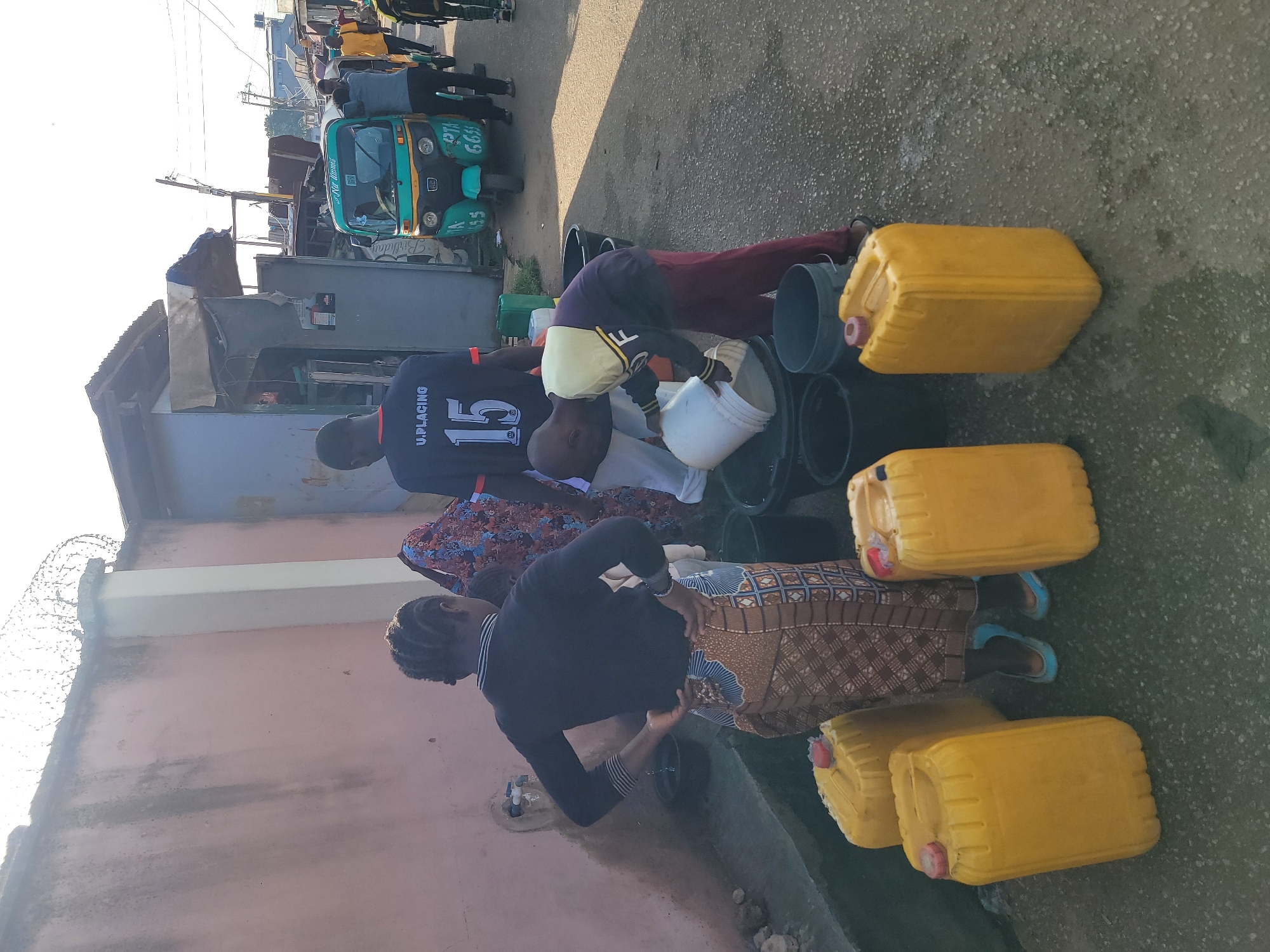 In Jos, Nigeria, this image captures a scene of community water collection. Several people are gathered around a water tap, filling large yellow jerrycans and black buckets. A young boy in a yellow hoodie is actively pouring water into a bucket, assisted by an adult. Another woman stands with her hands on her hips, seemingly waiting her turn. In the background, a teal and green auto-rickshaw is parked, and other individuals are visible further down the street, suggesting a busy urban environment. The scene takes place outdoors, near a wall, under a bright, likely morning, sky.