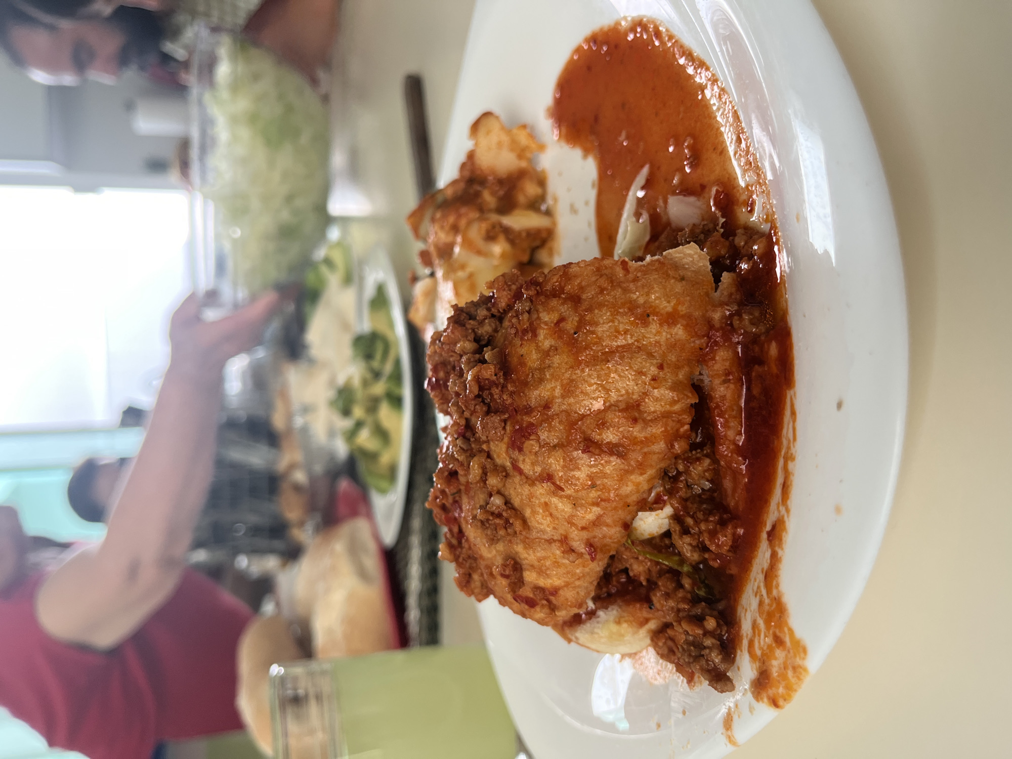 A white plate holds a pambazo, a Mexican sandwich, coated in red sauce. Some kind of white potato is on the plate. There is salad and bread on the table nearby. The location is possibly San Bernardino Tlaxcalancingo, Mexico, based on the sandwich type.