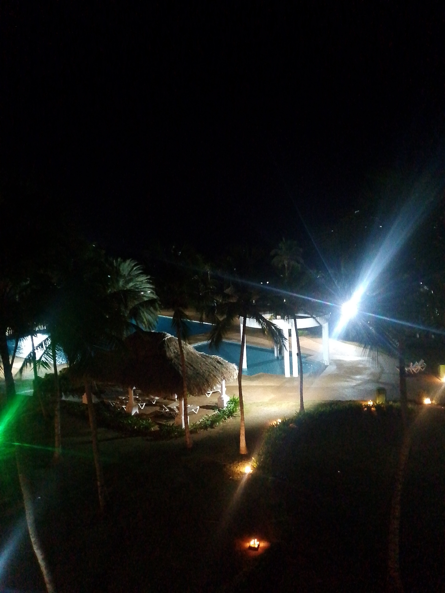 At night in Pedro González, Venezuela, a dimly lit outdoor scene reveals a swimming pool illuminated by bright lights, surrounded by palm trees and thatched umbrellas. The lighting casts a significant glare, obscuring details. Small, warm lights are sparsely placed along the ground, possibly lining a pathway. The overall atmosphere is serene despite the strong artificial lighting. No people are visible.