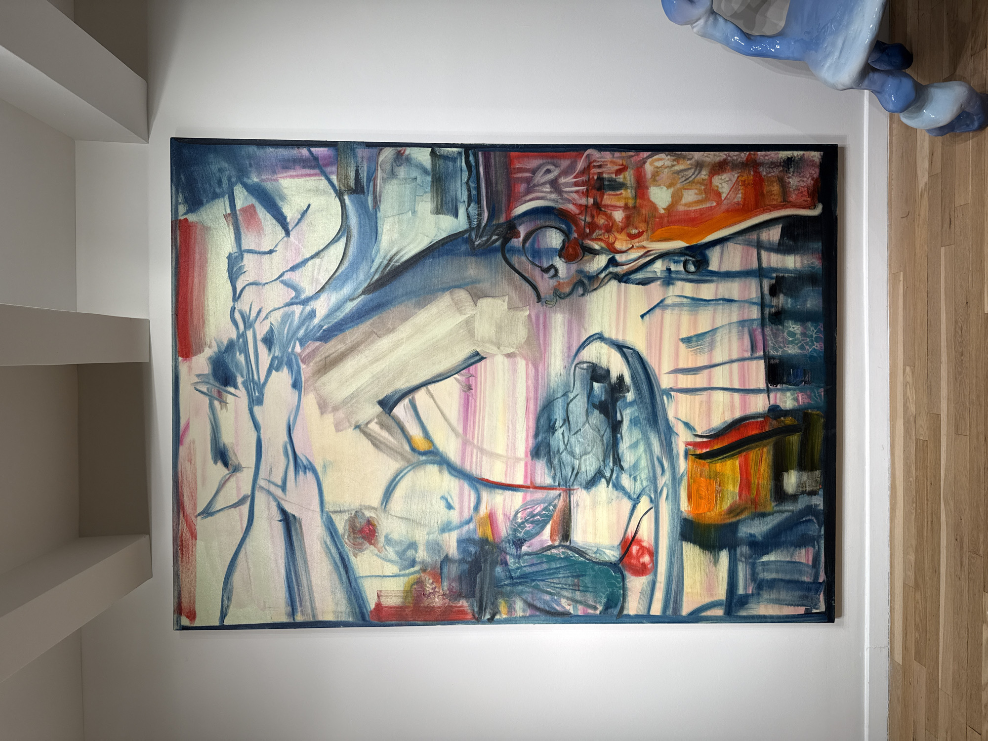 The image shows an abstract painting hanging on a white wall. The painting has a black frame and features a variety of colors, including blue, white, red, orange, and purple. The painting appears to be a mixture of brushstrokes and lines, with no clear recognizable objects or figures. The wall is smooth and white, and there are some white shelves visible on the left side of the image. To the right, a portion of a blue ceramic sculpture is visible, and further to the right, wooden flooring is present. The exact location in New York City, United States of America, cannot be determined from the image alone.