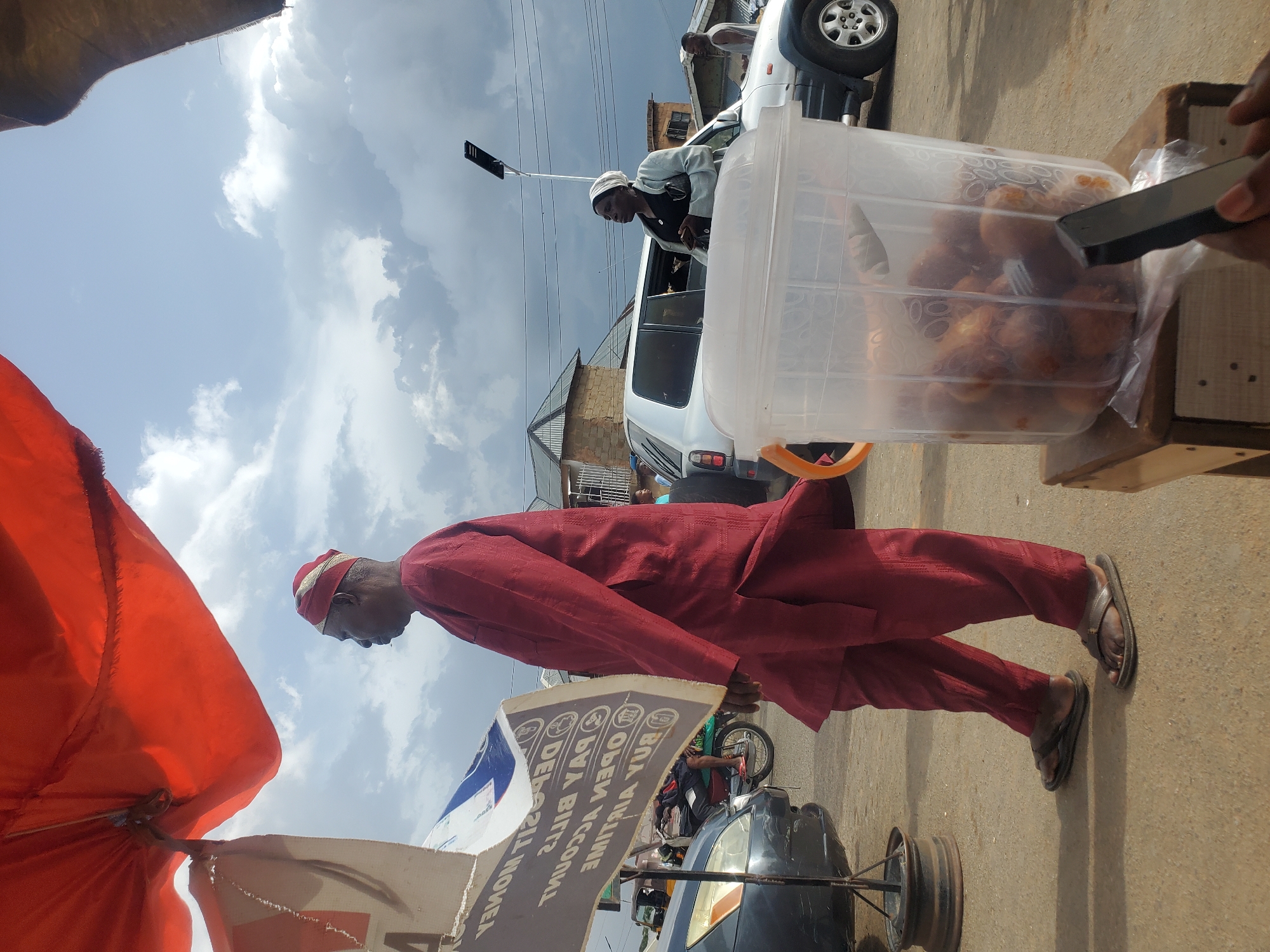 This image depicts a street scene in Gwong, Nigeria. The primary subject is a man walking in the foreground, dressed in a maroon traditional outfit and a matching cap. He is wearing dark flip-flops. Beside him, an orange umbrella is partially visible, suggesting an outdoor market or street vendor setup.
In the middle ground, a white SUV is parked. A person, possibly a woman, is leaning into the SUV from the driver's side, wearing a light-colored headscarf and a grey top. Another person, a man wearing a white cap and shirt, stands behind the SUV. A large, clear plastic container filled with fried snacks is prominently displayed on a wooden surface to the right of the SUV. A hand holding a smartphone is reaching towards the container.
In the background, other vehicles and buildings are visible, indicating an urban or semi-urban environment. The sky is partly cloudy, suggesting it could be late morning or afternoon. The overall atmosphere appears to be that of a busy street with commercial activity.
A sign or banner with text is visible in the lower left quadrant of the image, partially obscured by the umbrella. The visible text includes "DEPOSIT MONEY," "PAY BILLS," "BUY AIRTIME," and "OPEN ACCOUNT." These phrases suggest financial services are being offered at this location. The scene is well-lit, with shadows indicating daylight.