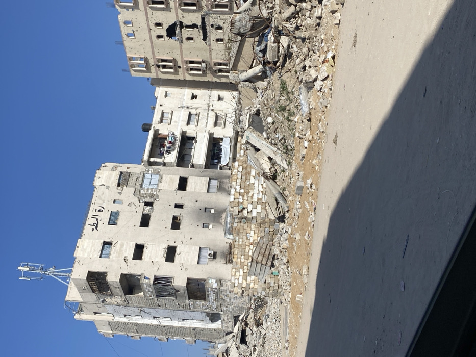 The image depicts a devastated urban landscape in the city of Khan Yunis, Palestinian Territory, under a clear blue sky. In the mid-ground, three multi-story residential buildings stand amidst extensive rubble.
The leftmost and central building, approximately 6-7 stories tall, features a light-colored facade heavily pockmarked by impacts. Multiple windows are empty, and sections of the exterior are damaged, revealing interior spaces. Black scorch marks are visible on the lower levels of this building. On its upper facade, Arabic script reading "امارة البطة" (Emirate of the Duck) is painted, and a cellular antenna tower is mounted on the roof. Balconies on the lower-right section of this structure show clothes hanging.
To the right, a slightly set-back, less damaged multi-story building, possibly 4-5 stories, is partially visible with laundry on its balconies and a black water tank on its roof. Further right, another 5-6 story building, also light-colored, shows significant structural damage including large, irregular breaches in its facade and numerous smaller impact marks. This building features distinctive arched window designs.
In the foreground, a large accumulation of demolition debris covers the ground, consisting of broken concrete slabs, twisted metal, and assorted rubble, including remnants of a wall constructed from white concrete blocks. The immediate foreground shows a sandy or dirt road surface, partially covered by a dark shadow extending from the lower left. No people are visible in the scene.