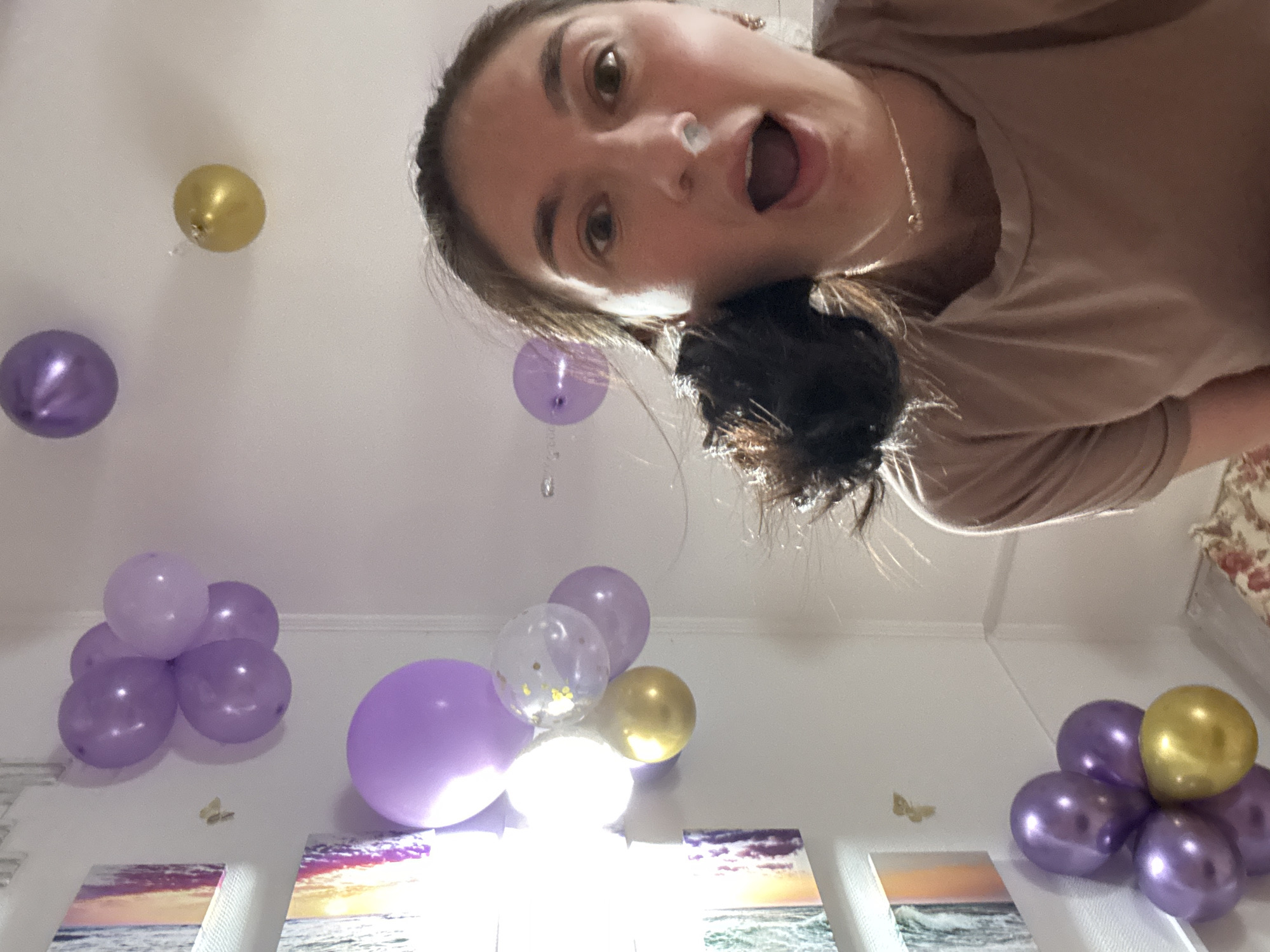 A woman with brown hair pulled back and wearing a tan shirt is holding a camera up and pointing down towards the room, causing a fish-eye lens effect. Her mouth is open in an exaggerated expression. The room is decorated with purple and gold balloons, some clustered together and some floating individually. There are three pictures hanging on the wall, depicting beach scenes. Small butterfly stickers are on the wall near the balloon clusters. The location is likely indoors, judging by the wall decor and the absence of outdoor elements, but the exact location within Sochi, Russia cannot be determined.