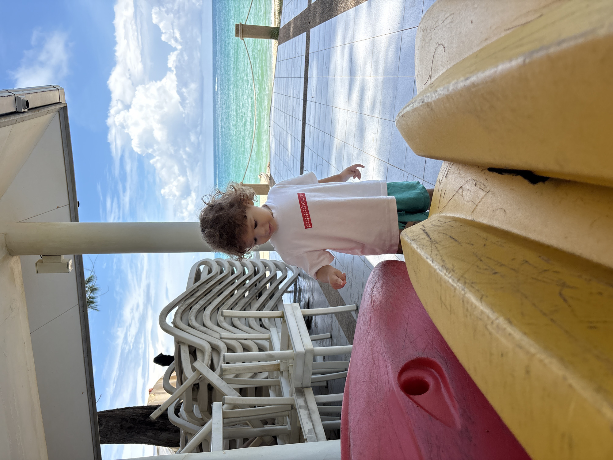 A young child with curly brown hair stands on a tiled patio near the ocean in Choeng Thale, Thailand. The child is wearing a white t-shirt with a red rectangular logo that reads "pappahappy" and teal shorts. In the foreground, the backs of two kayaks, one red and one yellow, are visible. Behind the child, a stack of white lounge chairs is piled high, and beyond that, a calm turquoise ocean stretches to the horizon under a blue sky with scattered white clouds. The scene appears to be at a resort or beachside location.