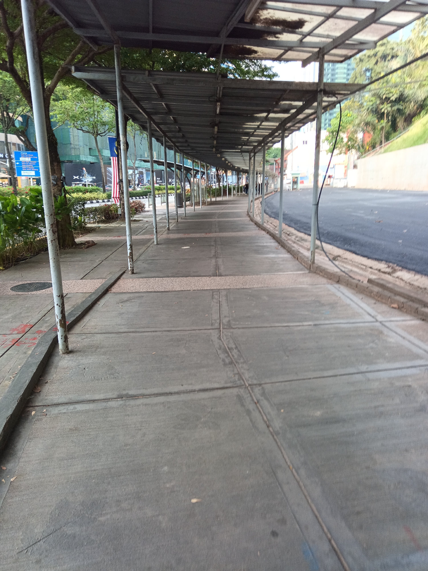 The image depicts a covered walkway along a city street in Kuala Lumpur, Malaysia. The walkway has a metal roof supported by a series of evenly spaced white poles. The ground of the walkway is paved with concrete slabs. To the left of the walkway is a sidewalk with some vegetation and trees. Behind the vegetation, buildings and a Malaysian flag are visible. To the right of the walkway is a dark asphalt road. The scene appears to be an outdoor urban environment.