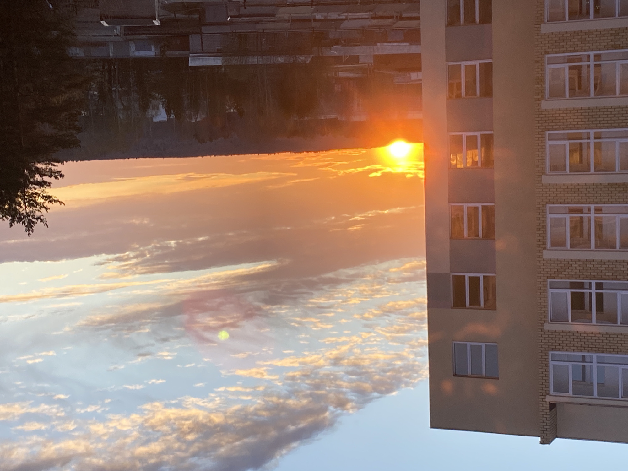 The image shows a sunset over what appears to be the city of Chaikovsky, Russia. The sun is positioned just above a treeline in the distance, radiating a bright orange glow across the sky. Below the sun is a layer of clouds tinged with gold. The sky transitions to a blue color lower in the frame, and some clouds are visible. A building is visible on the right. It is several stories tall with a mix of grey and brick facade. Multiple windows are visible. Some reflect the light from the setting sun. On the left edge of the frame, there is foliage. Above the tree line, an ambiguous shape is visible, but more information is needed to identify this.