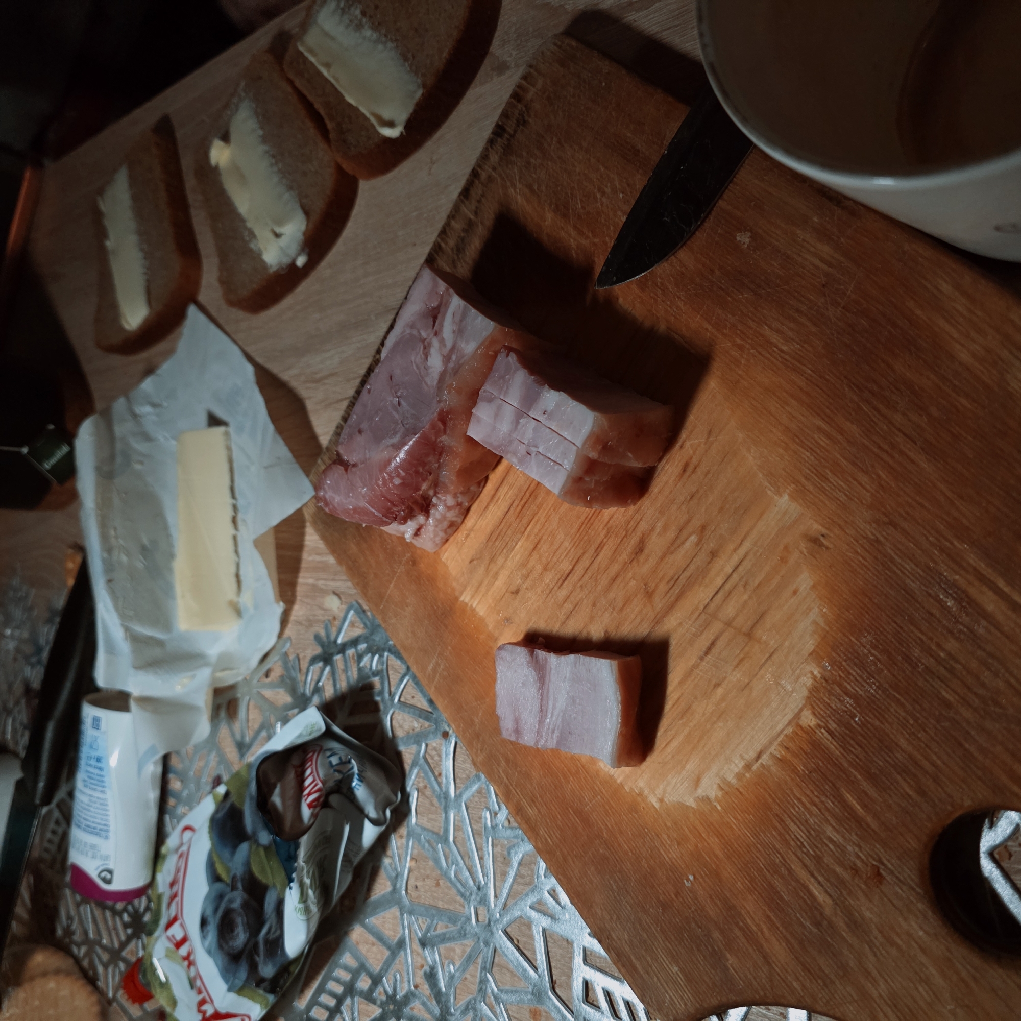 The scene depicts a tabletop view of food preparation in a domestic setting, likely a kitchen or dining area, within the city of Saint Petersburg, Russia.
A light-colored wooden cutting board, exhibiting knife marks and wear, occupies the central and lower portions of the frame. On this board, three pieces of raw pork fat with lean meat (salo) are arranged. One large, irregularly shaped piece, showing distinct layers of white fat and pinkish-red meat, is positioned towards the upper right of the board. Adjacent to it, to its lower left, is a smaller, cuboid piece of salo with horizontal score marks on its top surface. A third, thinner, rectangular slice of salo lies to the far left on the board. A dark-handled knife with a dark blade rests on the lower right portion of the cutting board.
To the upper right of the cutting board, three slices of dark bread are visible, each topped with a spread of butter. Above these bread slices, a block of butter is partially unwrapped from its white paper packaging. A white ceramic mug containing a light brown liquid is partially visible at the bottom right corner of the frame.
To the upper left, a metallic-patterned placemat with a geometric design is visible beneath a partially open package labeled "MAX & ELI" with "BLUEBERRY" clearly readable, suggesting it contains dried berries. A white and purple cylindrical container lies on its side behind this package. A green tea bag tag with "Greenfield" text is faintly visible in the upper central background. The lighting is localized, casting some shadows and highlighting the prepared items. No individuals are present in the image.