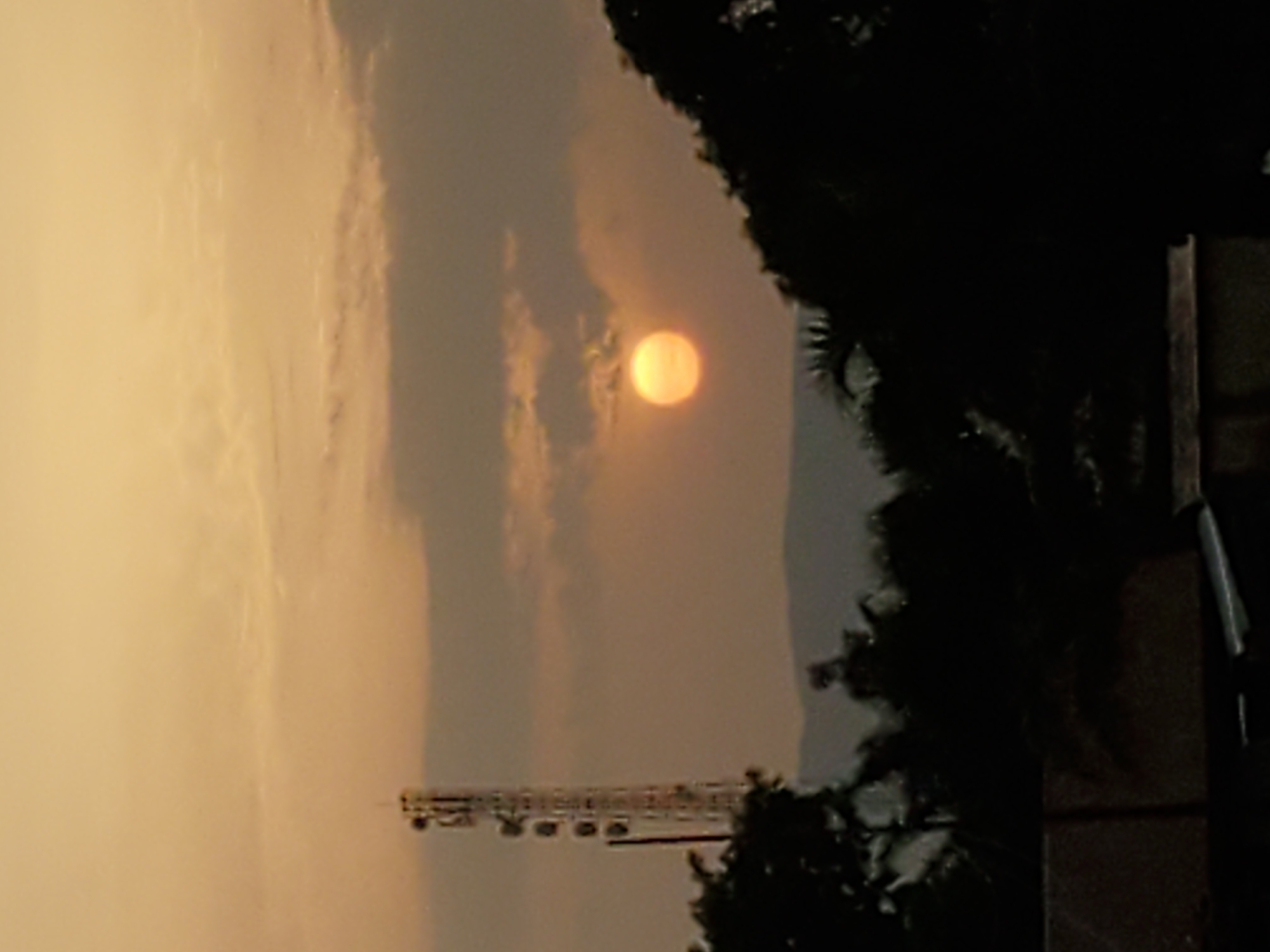The image depicts a sunset in Mararaba, Nigeria. A bright, hazy sun is positioned in the center of the frame. The sky is filled with yellow and gray clouds. Trees are silhouetted against the sky on the right side of the frame. An electrical support structure is visible near the bottom of the frame.