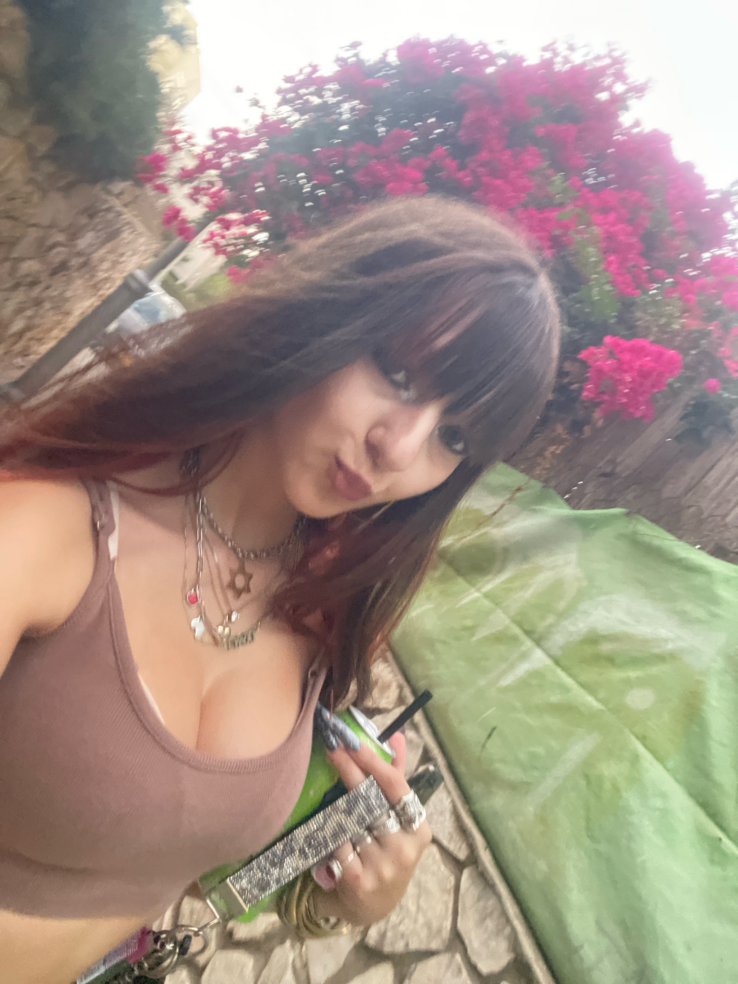 A young woman with long, dark brown hair and bangs is taking a selfie outdoors. She is wearing a brown tank top and multiple necklaces, including one with a Star of David pendant. Her left hand, adorned with several rings, holds a green cup with a black straw. Behind her, vibrant pink flowers bloom on a bush, contrasting with a stone wall on the left and a draped green object on the right. The scene appears to be in a residential area, possibly a garden or a street in Raanana, Israel, given the architectural style of the stone wall and the lush vegetation. The overall lighting suggests it is daytime, possibly overcast.