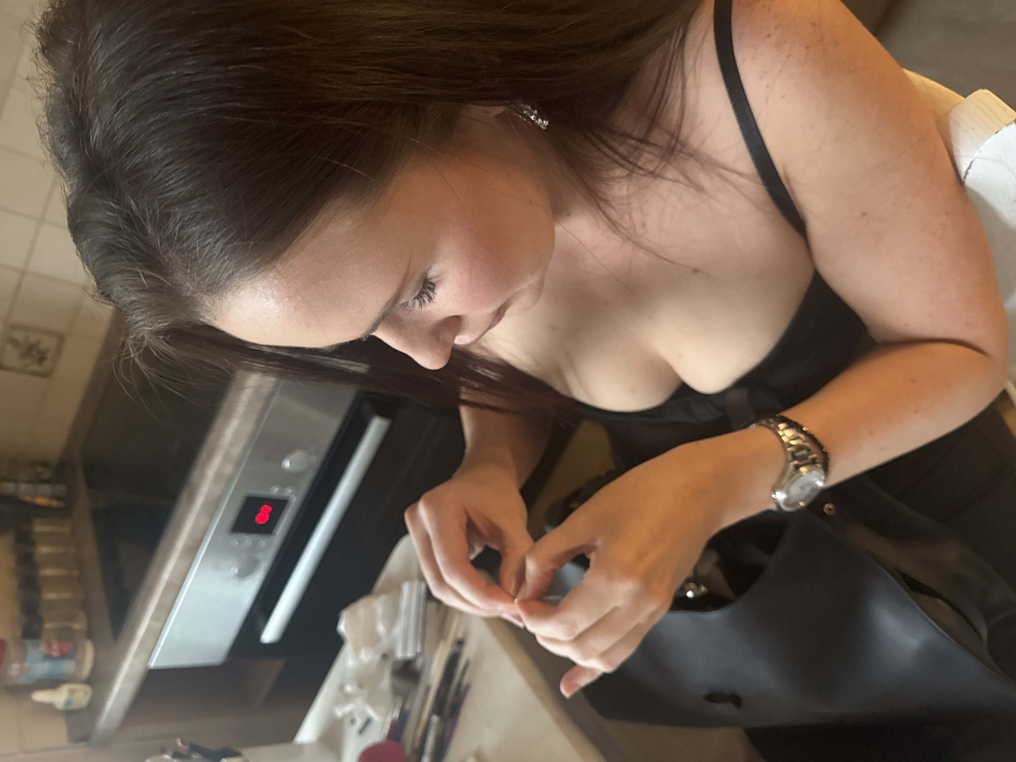 A woman with long dark hair is sitting in a room, possibly a kitchen, wearing a black top and a watch. She is looking down and appears to be fixing something with her hands. The background shows a stovetop, oven, and various items on a counter, including spice jars and makeup brushes. The lighting is natural, suggesting it could be daytime.