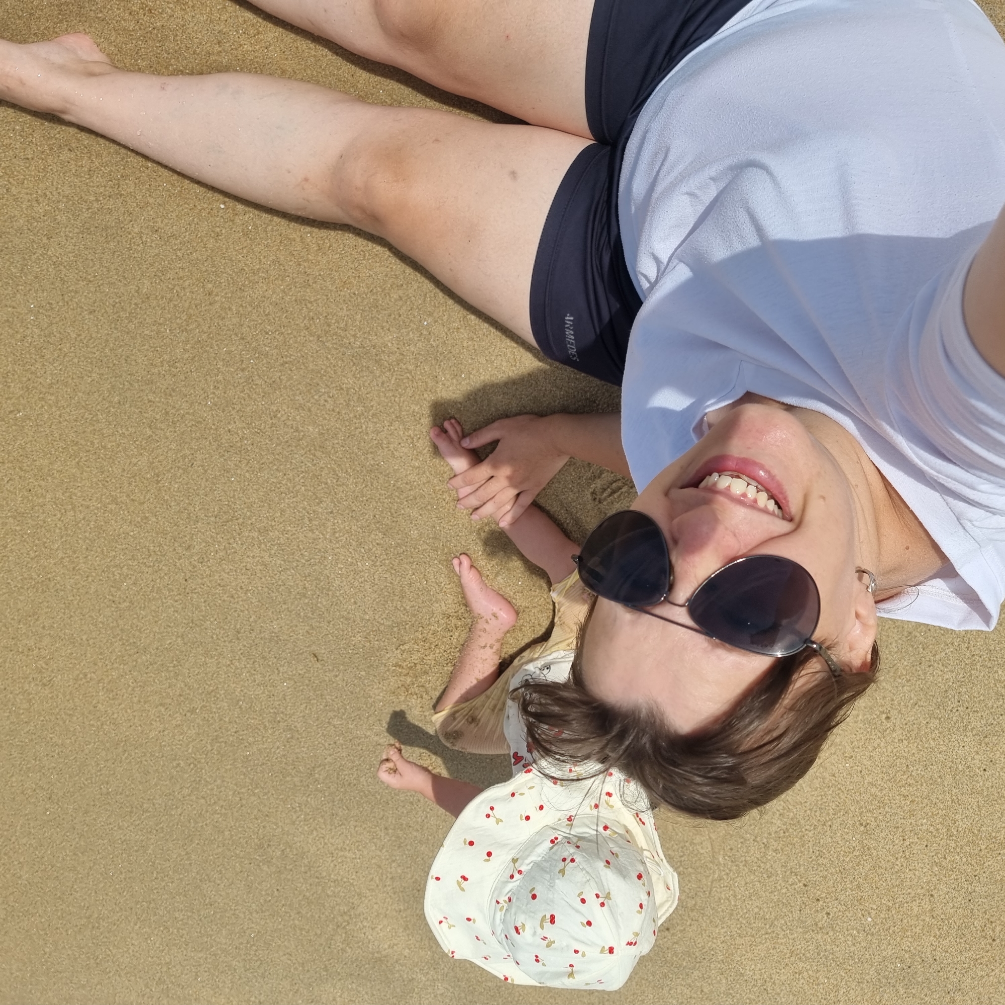 A woman and a child are on a sandy beach, likely in Gangneung-si, South Korea. The woman, in the foreground, is wearing sunglasses and a white t-shirt, and smiling at the camera. She is holding the child's hand. The child, further in the background, is wearing a sun hat with a cherry pattern and yellow shorts, and is interacting with the sand. The sand is light brown and appears to be wet in some areas. The lighting suggests it is a sunny day.