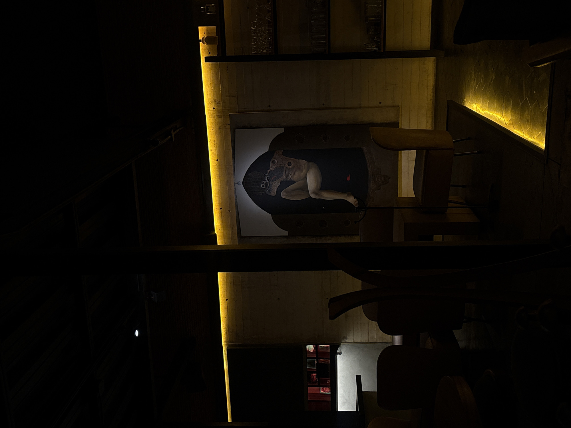 The image, taken in San Andrés Cholula, Mexico, captures a dimly lit interior with a focus on architectural elements and an unusual artwork. Vertical light strips illuminate concrete walls. The artwork depicts a nude human figure with exposed organs and bones, contained within an oval frame. The scene includes dark wooden furniture, possibly chairs and tables, and hints of additional interior spaces, some with windows displaying red hues. The lighting and composition create a somber, contemplative atmosphere.
