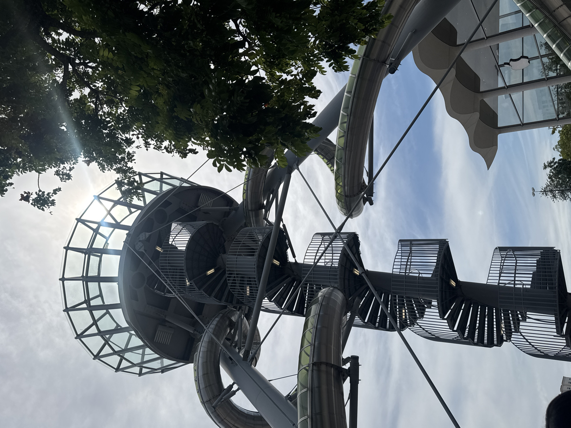 The image shows a spiral staircase and a glass-enclosed observation deck of a modern amusement park ride. The structure is made of metal and glass and is surrounded by lush green trees. In the background, a portion of a building with a distinctive wavy roof and an Apple logo is visible. The sky is partly cloudy with the sun shining brightly, creating lens flares. The scene appears to be outdoors, suggesting a daytime setting in a public space like an amusement park or recreational area. The location is identified as the city of Aventura, United States of America.