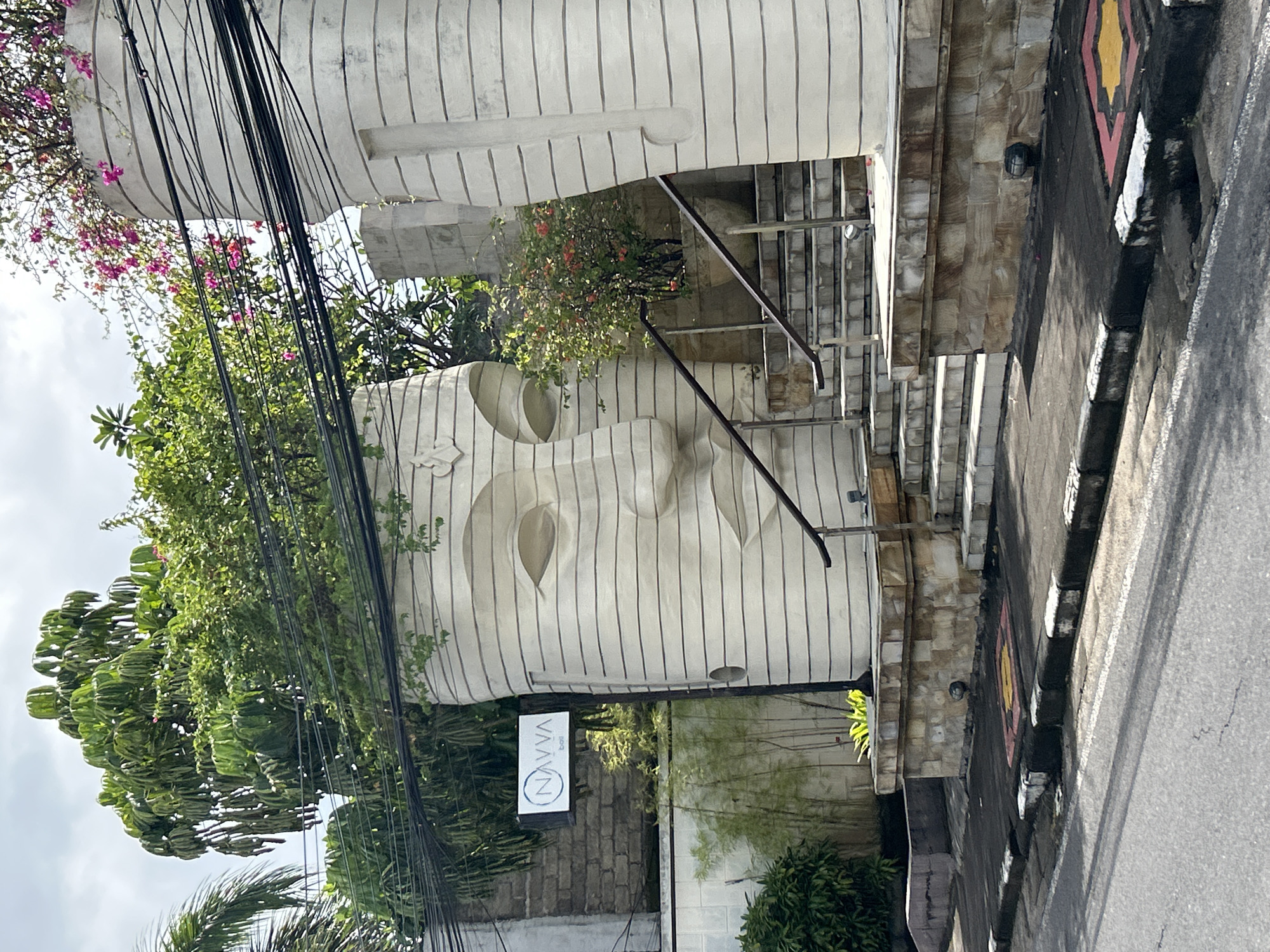 A large, sculpted head with stylized facial features forms part of a building facade. The head, constructed with linear striations, is located in Kuta Selatan, Indonesia. Plants grow from the top of the head, creating a natural crown of greenery. A sign with the word "NAVVA" is visible near the base of the head. A set of stairs leads up into the building. Power lines are in the foreground. A street and sidewalk are on the right side of the image.