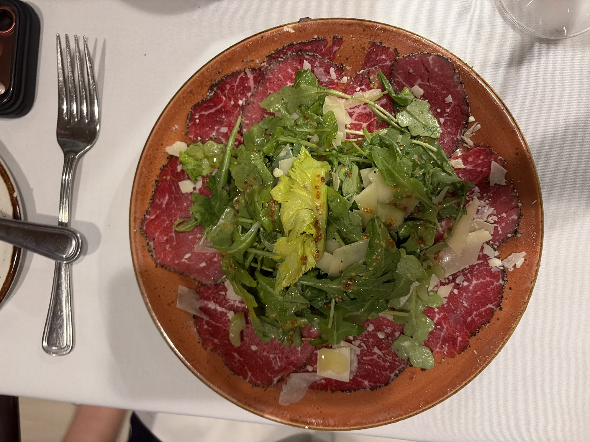 A high-angle shot captures a plate of beef carpaccio with a salad on a white tablecloth, accompanied by silverware. The carpaccio forms a ring around the plate, topped with a green salad featuring lettuce, arugula, and what appears to be a mustard seed dressing. Shaved Parmesan cheese is scattered across both the meat and the salad. A fork is positioned to the left of the plate, next to a partial view of a knife. A smartphone is also visible at the top left corner of the frame. The scene is likely set in a restaurant located in Key Biscayne, United States of America.