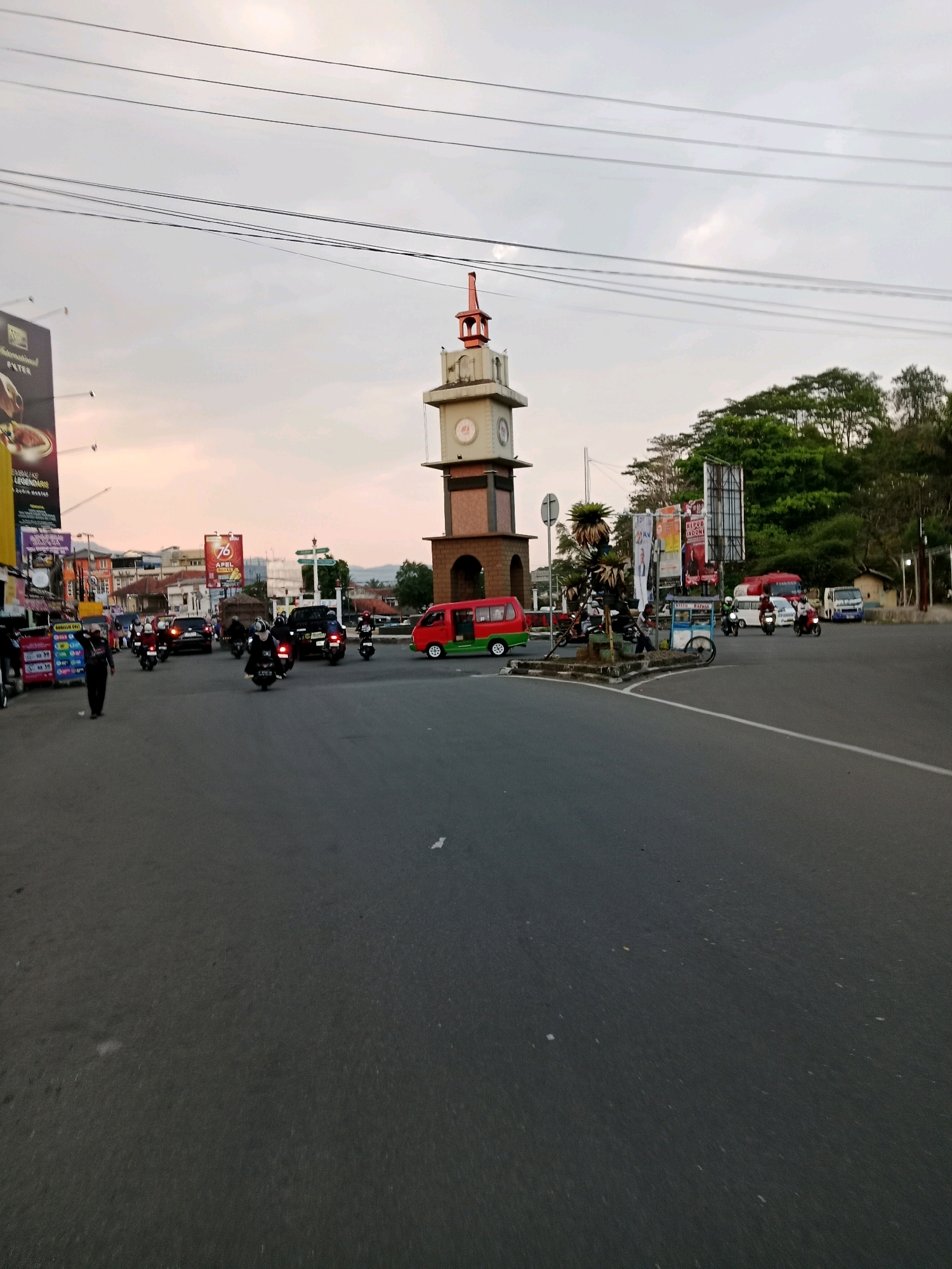 The image shows a street scene in Sirnagalih, Indonesia. A clock tower stands prominently in the background, with traffic and pedestrians present on the road. The sky is overcast, suggesting it might be early morning or late afternoon. The road is paved and appears to be a busy intersection with several vehicles, including cars and motorcycles, moving in different directions. A red and green van is visible, along with a water tank truck. Several people are on foot, some appearing to be crossing the street or walking on the sidewalk. Billboards and advertisements are displayed along the sides of the road, contributing to the urban environment. The overall impression is a typical day in a bustling Indonesian city.