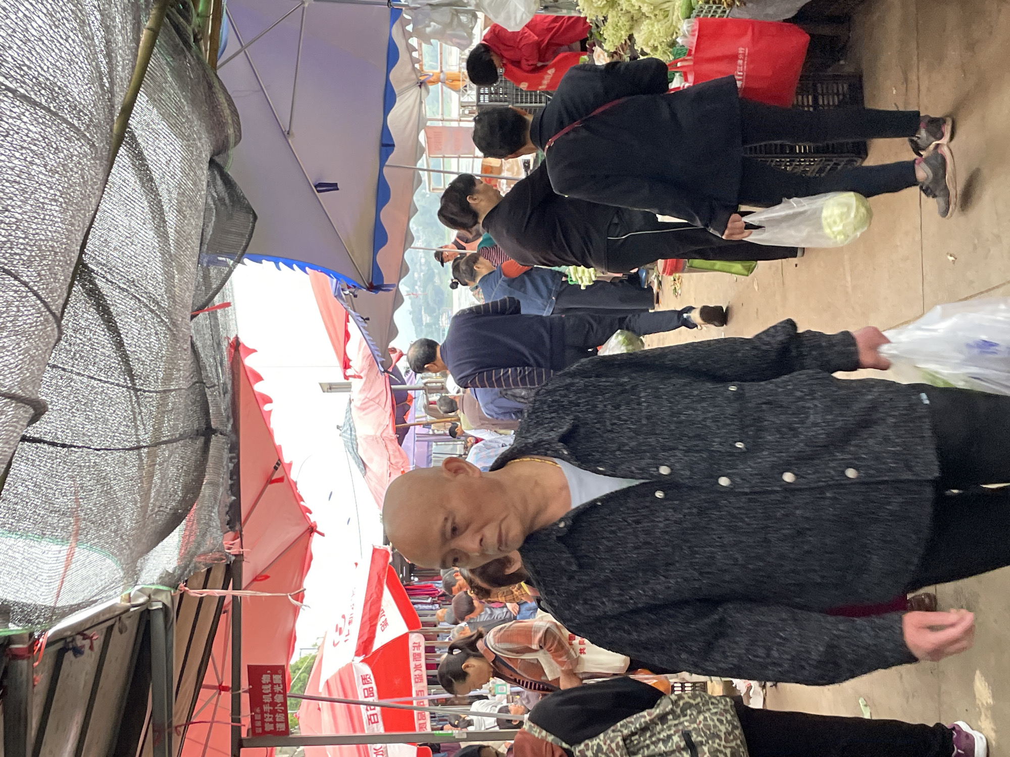 This outdoor scene depicts a crowded marketplace with numerous individuals shopping. The marketplace features stalls covered by a mix of black netting, red and white striped awnings, and blue-trimmed umbrellas. Shoppers of various ages are present, dressed in casual attire. One man in the foreground wears a gray speckled jacket and appears to be holding a plastic bag. Other shoppers are seen carrying produce in plastic bags and baskets. The market setting suggests a location in Jiangjin, China.