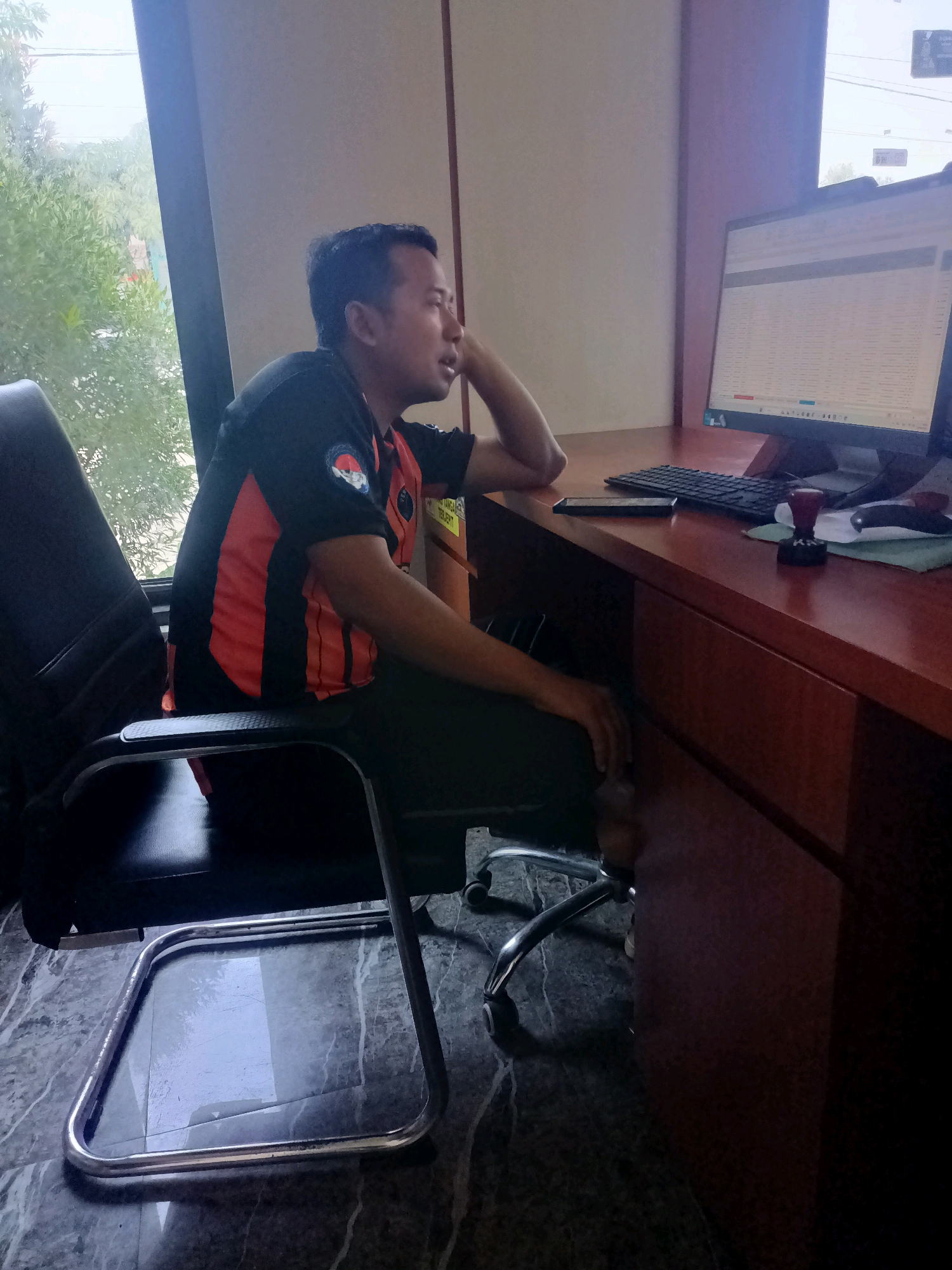 A man in a black and orange shirt sits in an office chair at a wooden desk, looking towards a computer monitor. The office appears to be located in Sumber Bendo, Indonesia, as indicated by the surrounding vegetation visible through the window. The man's right arm is propped on the back of the chair, and his hand rests on his head. The desk is cluttered with a keyboard, a smartphone, a rubber stamp, and papers. The floor is tiled with a marble pattern. The overall impression is that of a typical office environment.