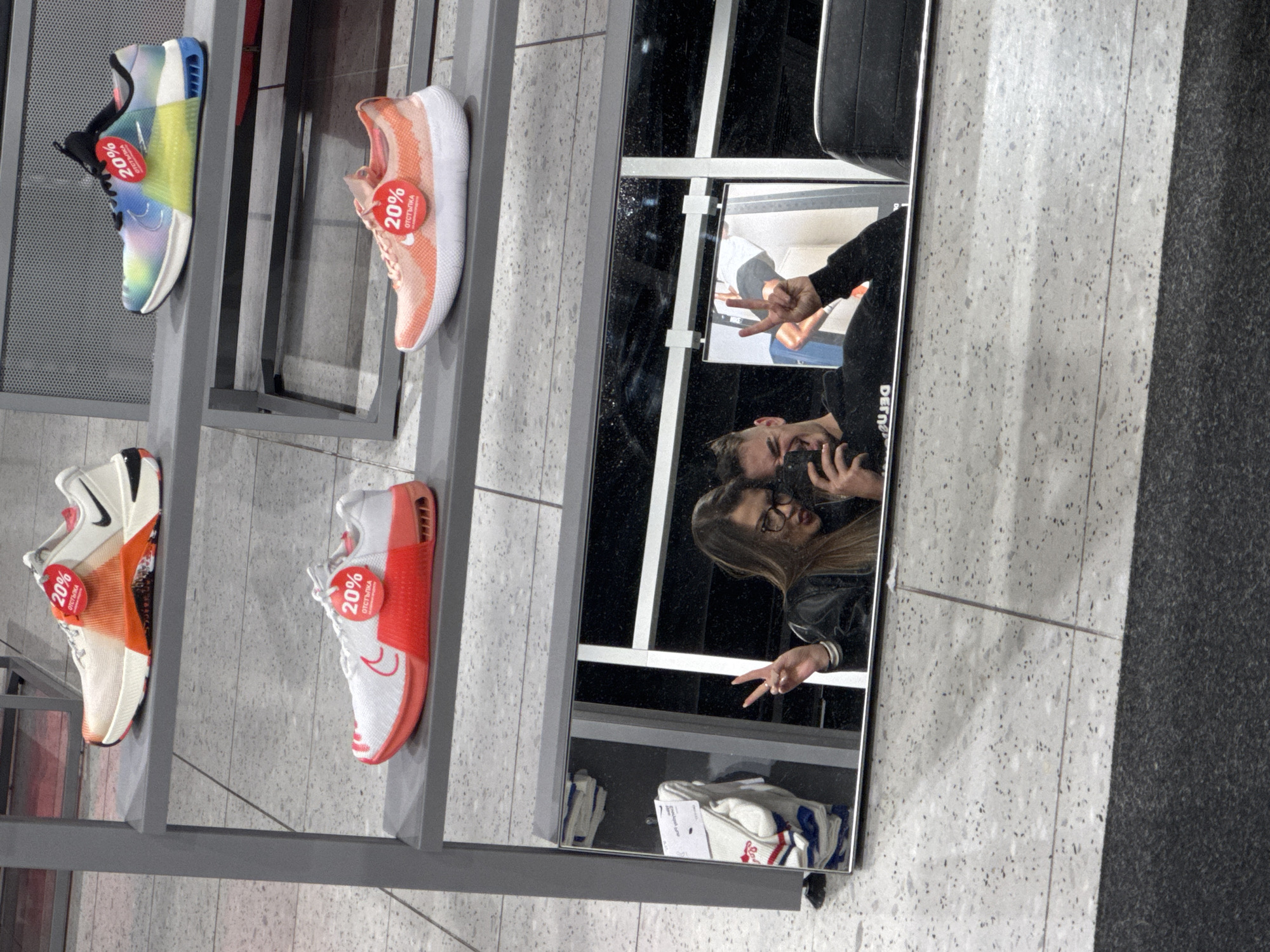 The image is taken inside a store, possibly in Sofia, Bulgaria, as suggested by the Russian text (отстъпка) on discount tags. A gray metal shelving unit displays four pairs of running shoes, each marked with a 20% discount tag. A large mirror adjacent to the shelving unit reflects a couple taking a selfie. The man is holding the phone, while the woman makes a peace sign. Clothes are also reflected in the mirror below the couple. The floor is a mottled gray and white tile, with a patch of dark gray, possibly a rug, in the right corner.