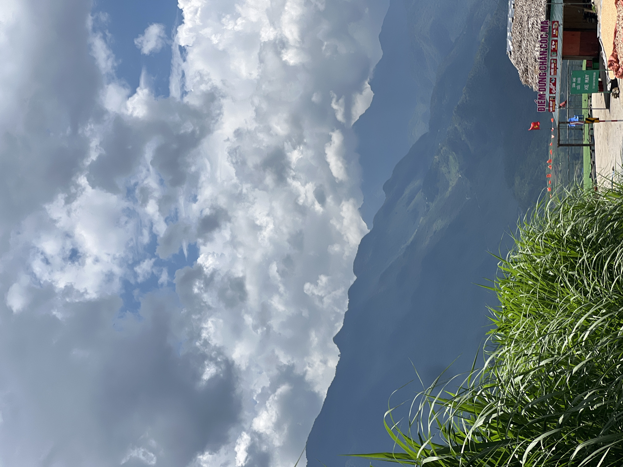 This image captures a scenic view in Lùng Tám Commune, Vietnam, characterized by lush green vegetation in the foreground, dramatic mountain ranges in the mid-ground, and a cloudy sky above. In the lower right corner of the image, a small building with a thatched roof is visible, featuring Vietnamese signage that reads "ĐIỂM DỪNG CHÂN CỐC MA" and other text that is partially obscured. A red flag is displayed on a pole in front of the building. A sign with green text on a white background, possibly indicating a parking area for motorcycles, is also present. The environment is mountainous and rural, with the presence of a structure that appears to be a rest stop or a small commercial establishment catering to travelers. The overall atmosphere is natural and serene, with a hint of human presence through the roadside building.