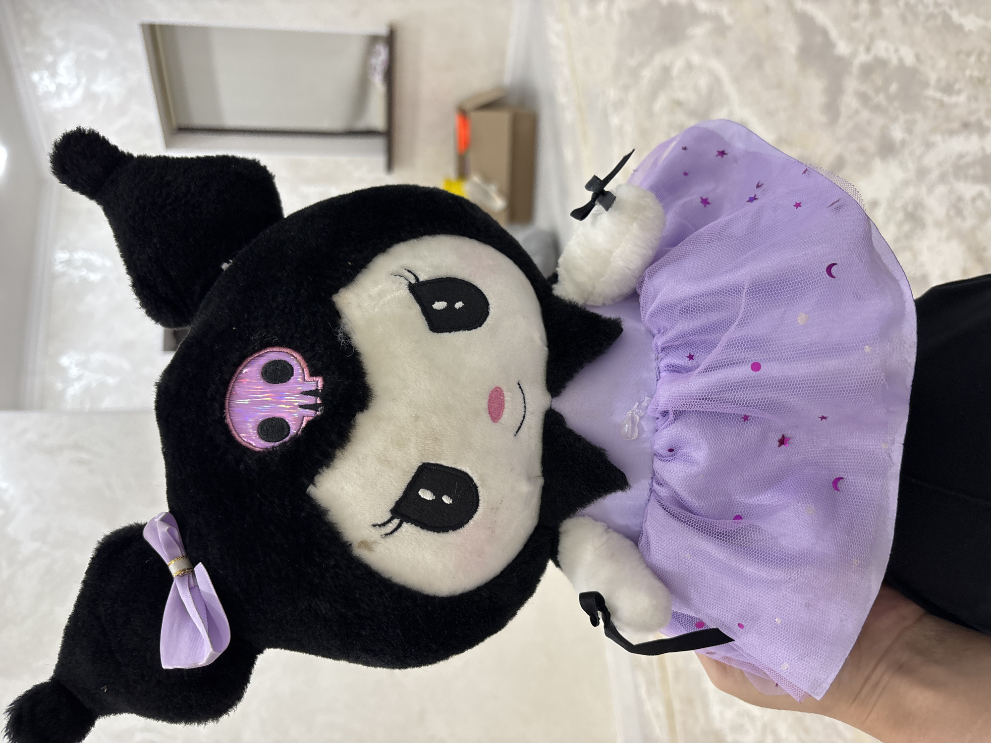 A plush toy, resembling the character Kuromi from Sanrio, is held in a person's hand. The toy is predominantly black and white, with large black ears, a black hood adorned with a pink skull emblem, and large black eyes with white pupils. It wears a black collar and a light purple tutu dress decorated with pink glittery moons and stars. Small black bows are attached to the toy's wrists. The background appears to be an indoor setting, possibly a room in a residential building in the city of Жамбыл, Kazakhstan, with textured walls and a slightly out-of-focus view of a doorway and a cardboard box in the distance.