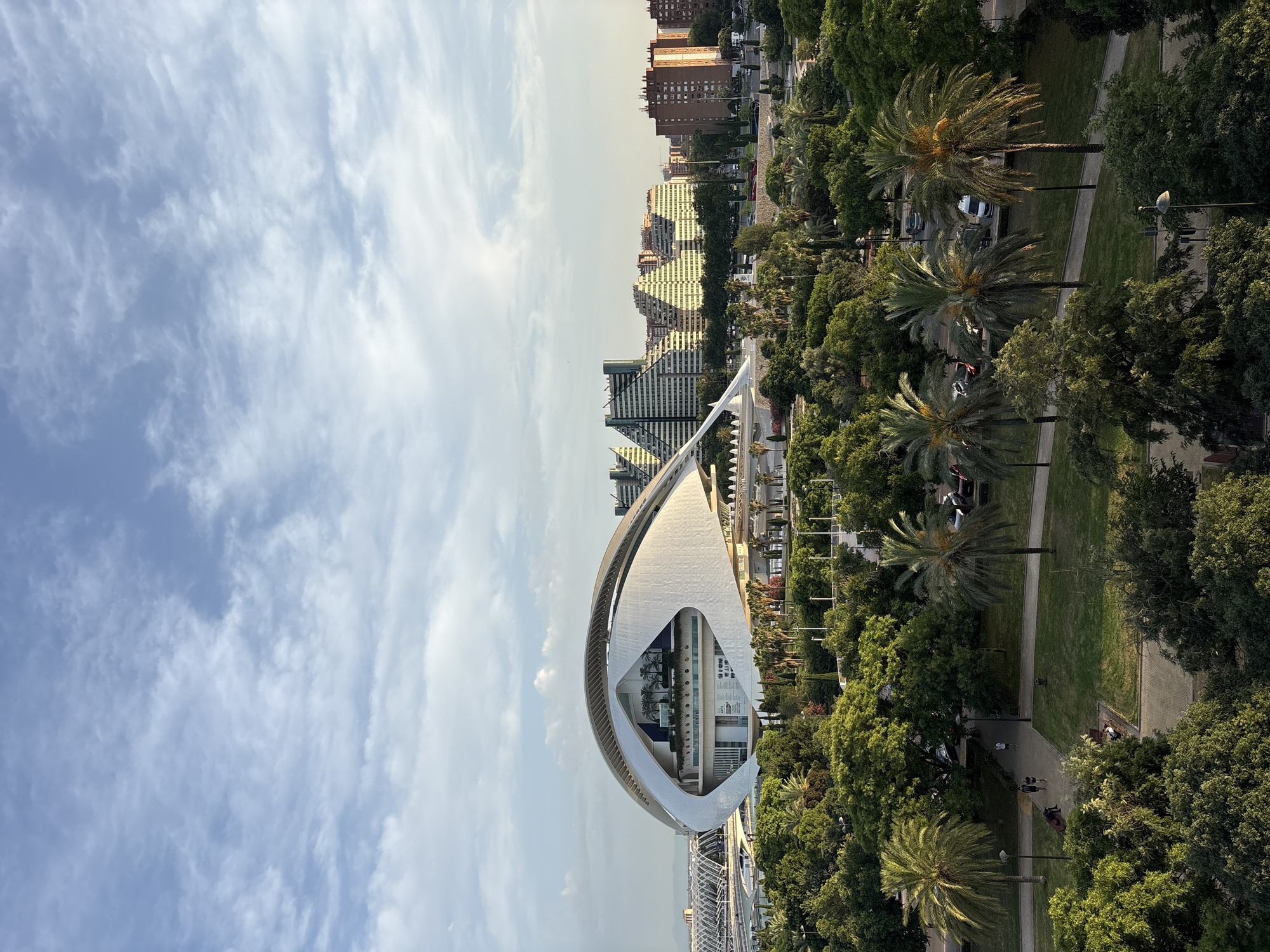This is a high-angle, exterior, medium shot of the City of Arts and Sciences in Valencia, Spain. The iconic L'Hemisfèric building dominates the center of the frame, its distinct "eye" shape creating a dramatic architectural feature. The building is white and curved, with a large opening revealing interior levels with palm trees. In the background, a cluster of modern, stepped apartment buildings with a repetitive, triangular facade are visible, along with other structures and a distant bridge.
The foreground is filled with lush green trees, including several mature palm trees with their fronds reaching towards the sky. A park area with paved pathways and grassy lawns is present in the middle ground. Several people are casually walking and sitting in the park. Cars are parked along a road in the distance, partially obscured by trees. The sky above is a pale blue with scattered, wispy white clouds, suggesting it is either early morning or late afternoon. The overall scene depicts a modern urban park with striking contemporary architecture.