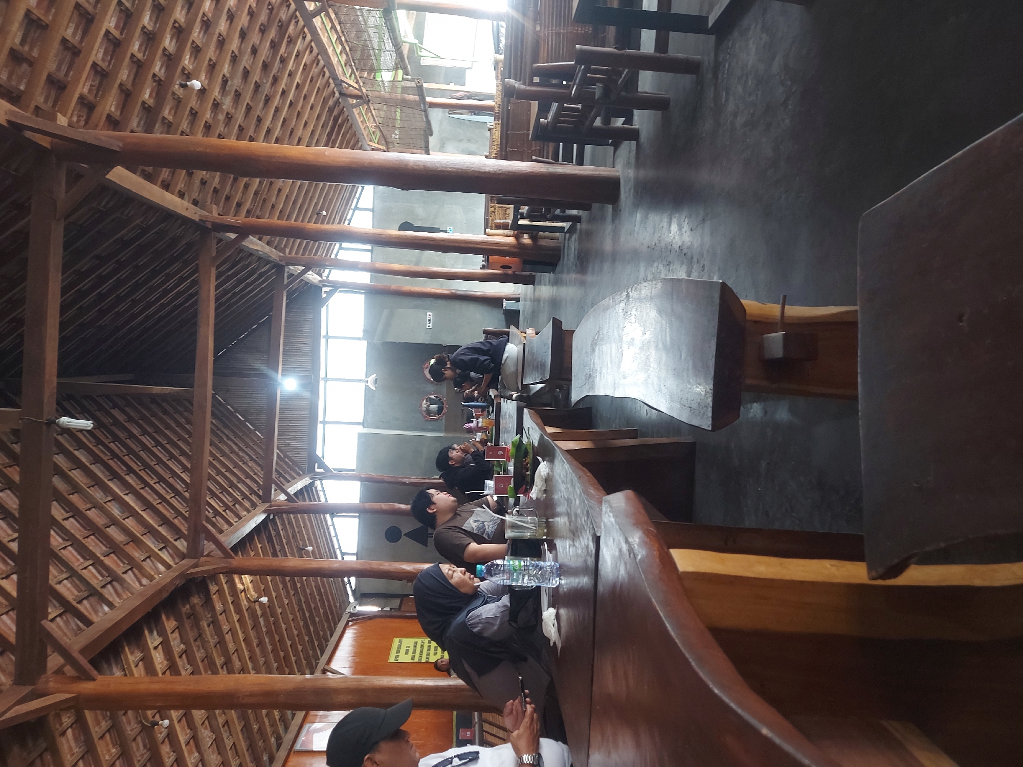 The image shows the interior of a restaurant with a natural, open-air design. The structure features a thatched roof supported by thick wooden pillars, creating a rustic and spacious atmosphere. Several wooden tables and benches are visible, arranged to accommodate diners. In the foreground, a long, curved wooden table is prominent, with a few people seated at it. One person in the foreground is wearing a white shirt and a black cap and is looking down at a mobile phone. Next to them, a woman in a dark hijab is seated, and in front of her is a bottle of water and a glass. Further down the table, other individuals are also seated, with food and drinks on the table. In the background, more seating areas and tables are visible, with some people in the distance. The lighting suggests it is daytime, with natural light entering from the open sides and through windows. The floor is a dark, concrete material. The overall impression is of a casual dining establishment with a focus on natural materials and an open ambiance. There is no indication of the specific location within Kricak, Indonesia, beyond it being an interior scene of a restaurant.