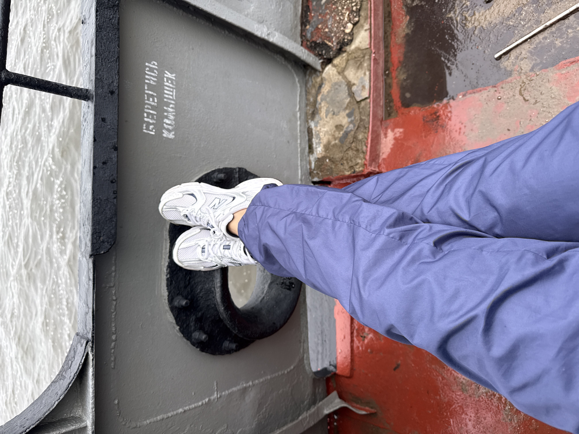 The image shows a person's legs and feet in blue pants and white sneakers, crossed and resting on the deck of a vessel. The person is positioned near a circular opening in the hull and a railing. In the background, there is choppy water. Stenciled white text on the hull reads "БЕРЕГИСЬ КОЛЫШЕК" (Beware of the bumps). The scene is outdoors, likely on a boat or pier, with a backdrop of gray, overcast skies and a body of water. The overall impression is one of casual relaxation or waiting on the deck of a ship.