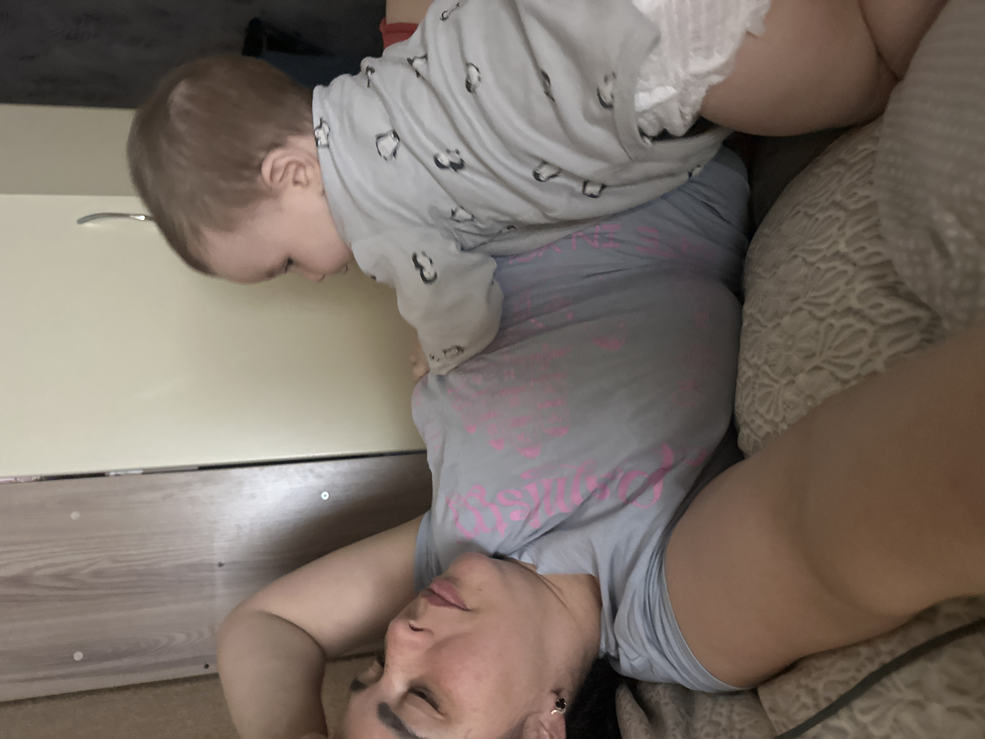 A photograph depicts a woman lying on her back with a child perched on her stomach. The woman is wearing a gray t-shirt with pink lettering, and her eyes are closed. The child is wearing a gray onesie with a penguin print. The background includes a headboard of a bed with a white wall behind it. The location is Unknown, Unknown.