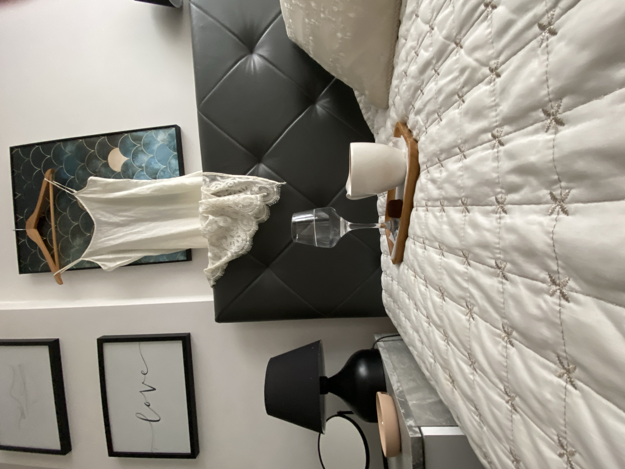 A photograph taken indoors shows a bedroom. There is a bed with a gray leather headboard. The bedspread is white with a pattern of embroidered crosses. There is a white pillow on the bed. A small wooden tray on the bed holds a white coffee cup and a wine glass. A white nightgown hangs on a wooden hanger on the wall next to a framed image of fish scales. Below that is a framed print of the word "love" in script and another framed print. A black lamp sits on the nightstand. The location of the photograph is unknown and the city of Budva, Montenegro cannot be confirmed.