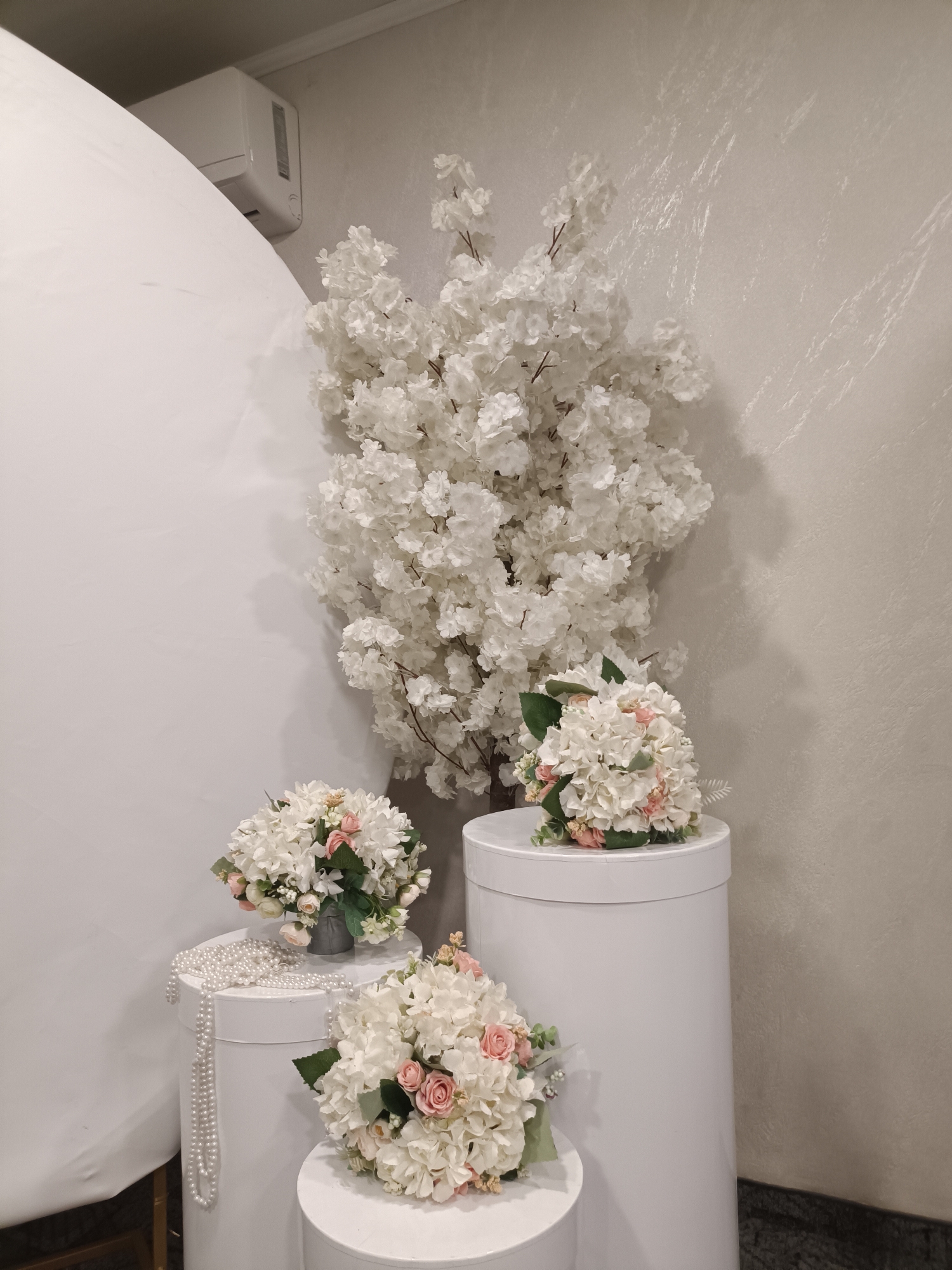 This image captures an elegant indoor decorative setup, likely within an event venue or studio in Plovdiv, Bulgaria. The central focus is a large, artificial white cherry blossom tree or bush, densely covered in delicate white flowers, standing against a light-colored, subtly textured wall.
Arranged in the foreground are three bouquets of artificial flowers, primarily composed of white hydrangeas with accents of soft pink roses and green foliage. These bouquets are displayed on white cylindrical pedestals of different heights. One bouquet rests on the tallest pedestal, another is presented in a small silver-grey vase on a medium-height pedestal, and the third is placed on the lowest pedestal. A long, gracefully draped strand of white pearl-like beads adds a touch of classic elegance, resting on one of the pedestals.
In the upper left corner, an air conditioning unit is mounted high on the wall, and a large, curved white panel or backdrop occupies the left side of the frame. The floor, visible at the bottom right, appears to be dark granite or marble.
The overall aesthetic, dominated by white and soft pink tones, evokes a sense of purity, sophistication, and celebration. While no specific activity is shown, the arrangement strongly suggests preparations for an event such as a wedding, bridal shower, or a formal gathering. No people are visible, and there is no discernible text within the image.