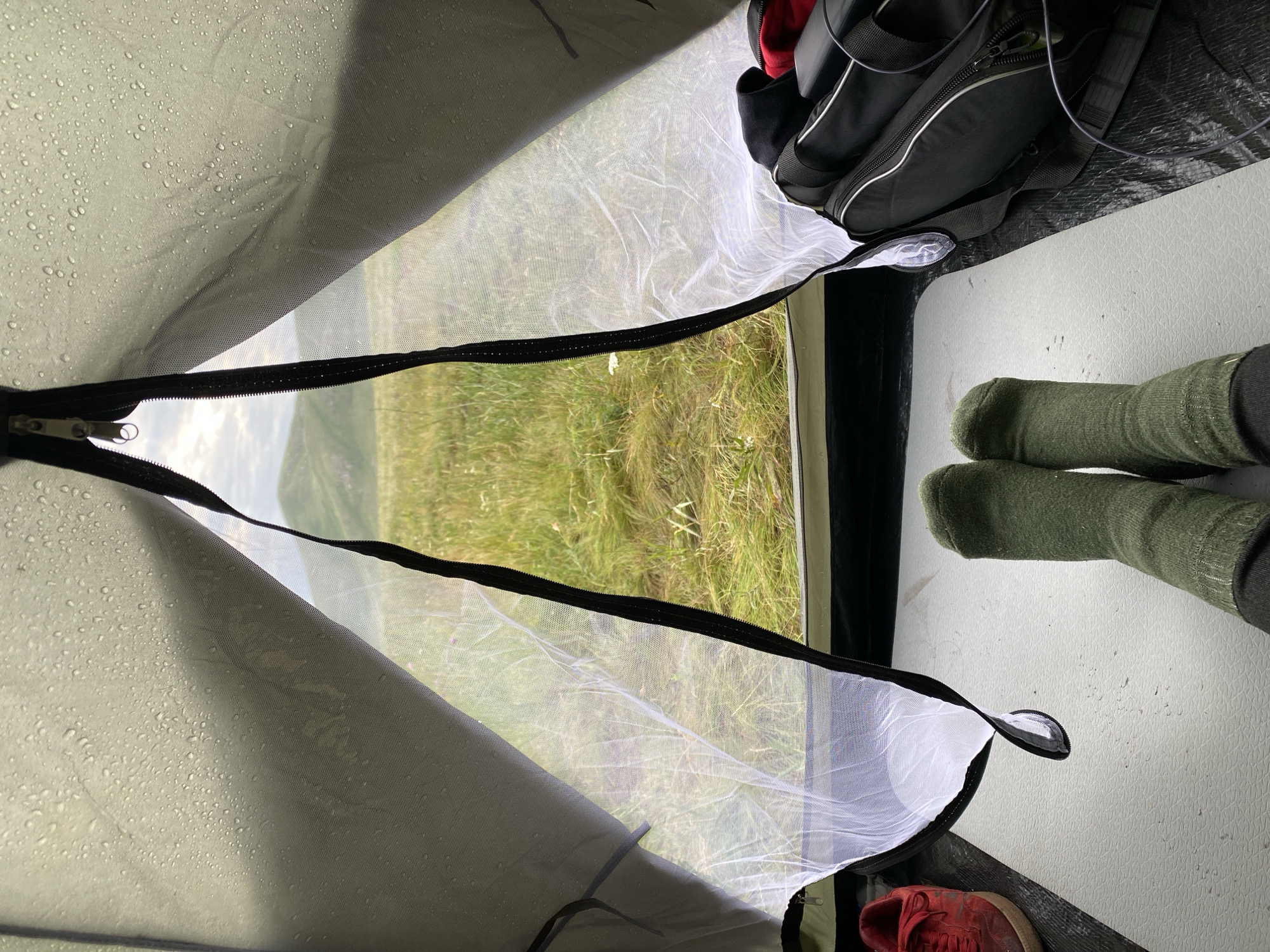 The image is taken from inside a tent, looking out at a landscape. The tent fabric, likely waterproof, shows numerous water droplets on its exterior. The tent opening is partially zipped, with a mesh layer visible. Outside, a grassy, undulating terrain with hills is visible under a cloudy sky.
In the foreground, inside the tent, a person's legs are visible, clad in dark green socks, resting on a light-colored mat. To the left, a red sneaker is partially visible. To the right, a black duffel bag with silver zippers and straps is placed on the tent floor, along with some black cables.
The overall scene suggests a person camping outdoors, possibly in a rural or mountainous area, with the weather being damp. The location is stated to be in the city of Абзаново, Russia, implying this is a view from a campsite in that region.